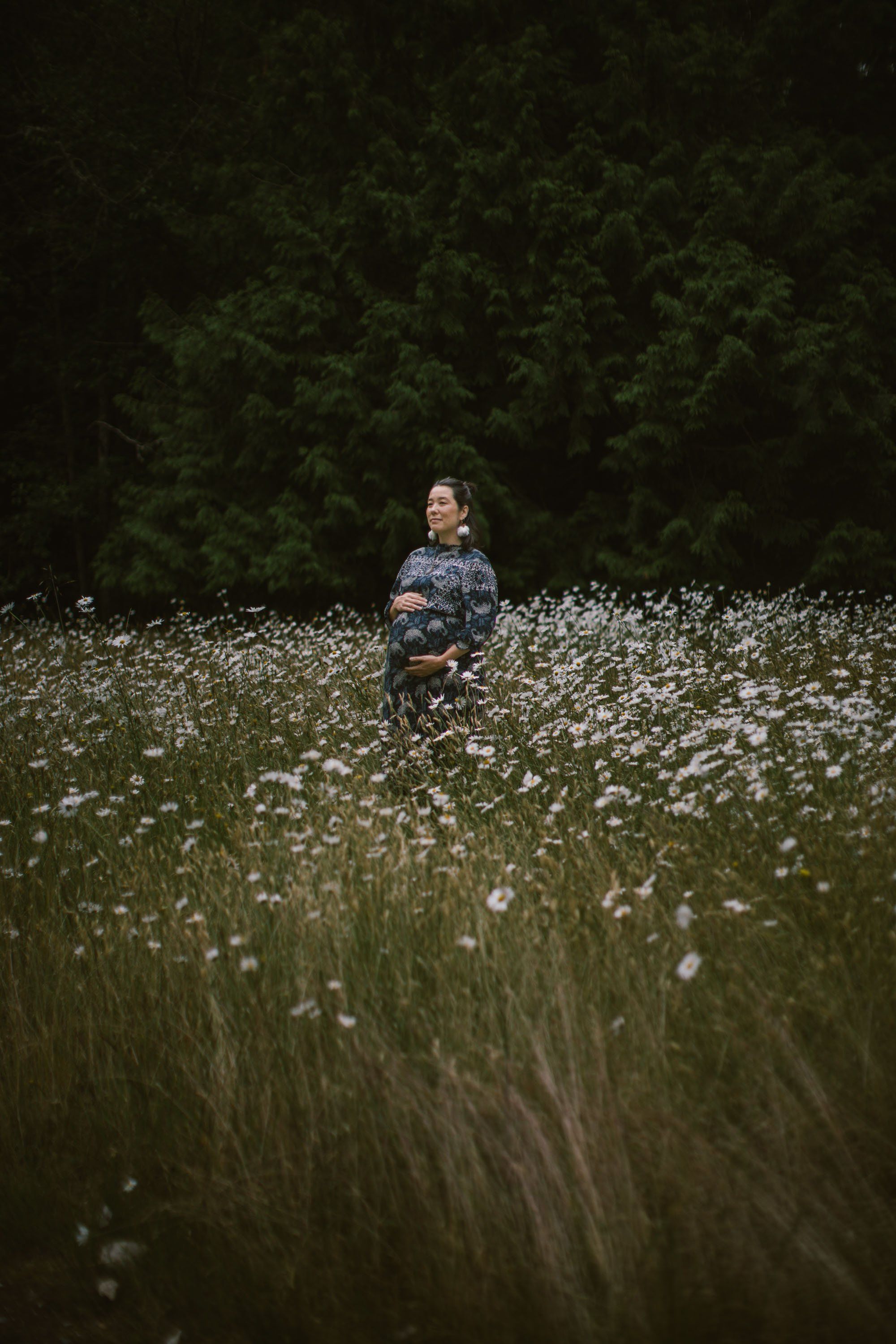 Pregnant woman in denim jacket standing in a field of white wildflowers with dark forest behind her