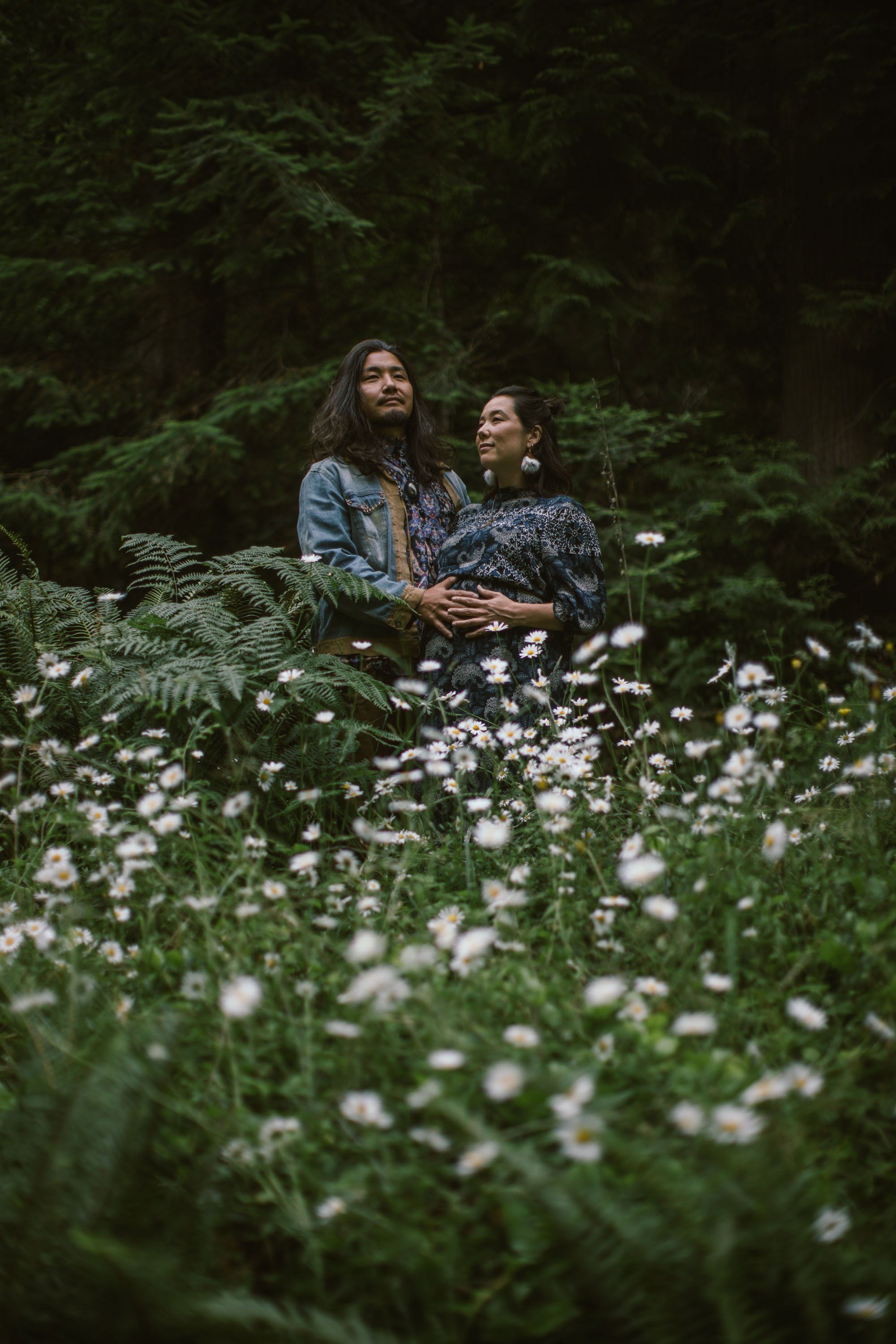 Expectant couple in denim amid a wildflower meadow, surrounded by dark forest, gazing upward together in golden light