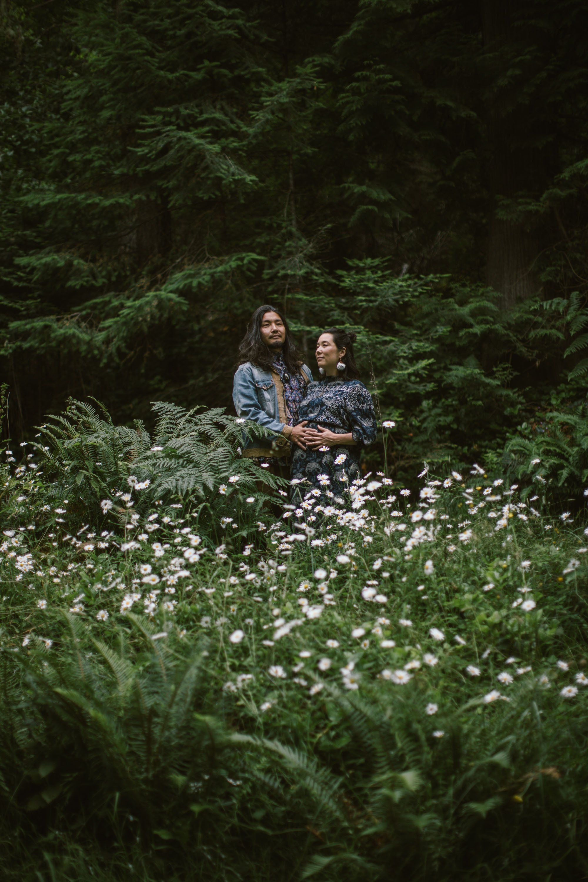 Two people nestled in a wildflower meadow surrounded by dense forest, soft natural light filtering through evergreen trees