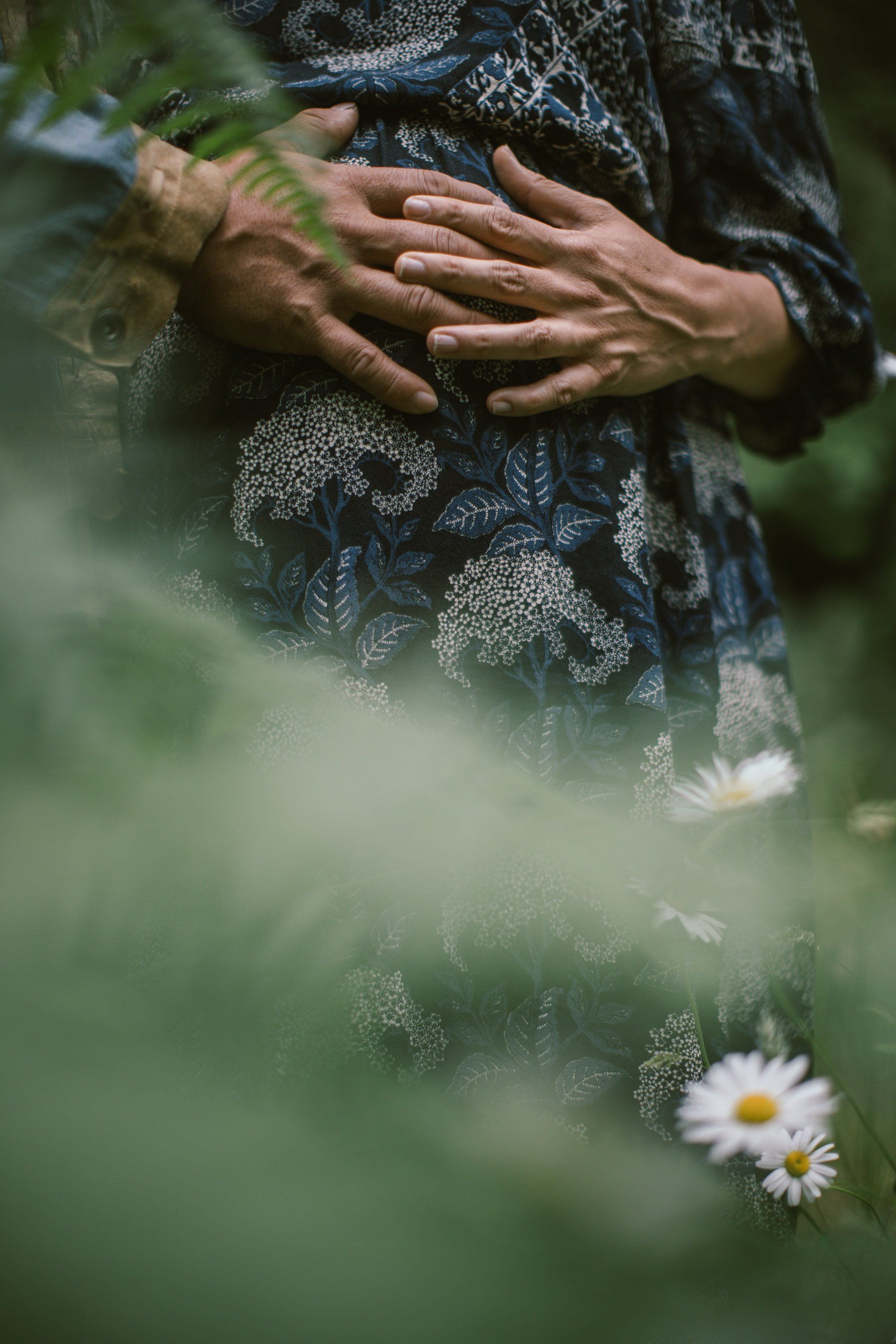 Hands cradling an expectant belly through sheer fabric, wildflowers soft-focused in foreground, intimate maternity moment in garden light