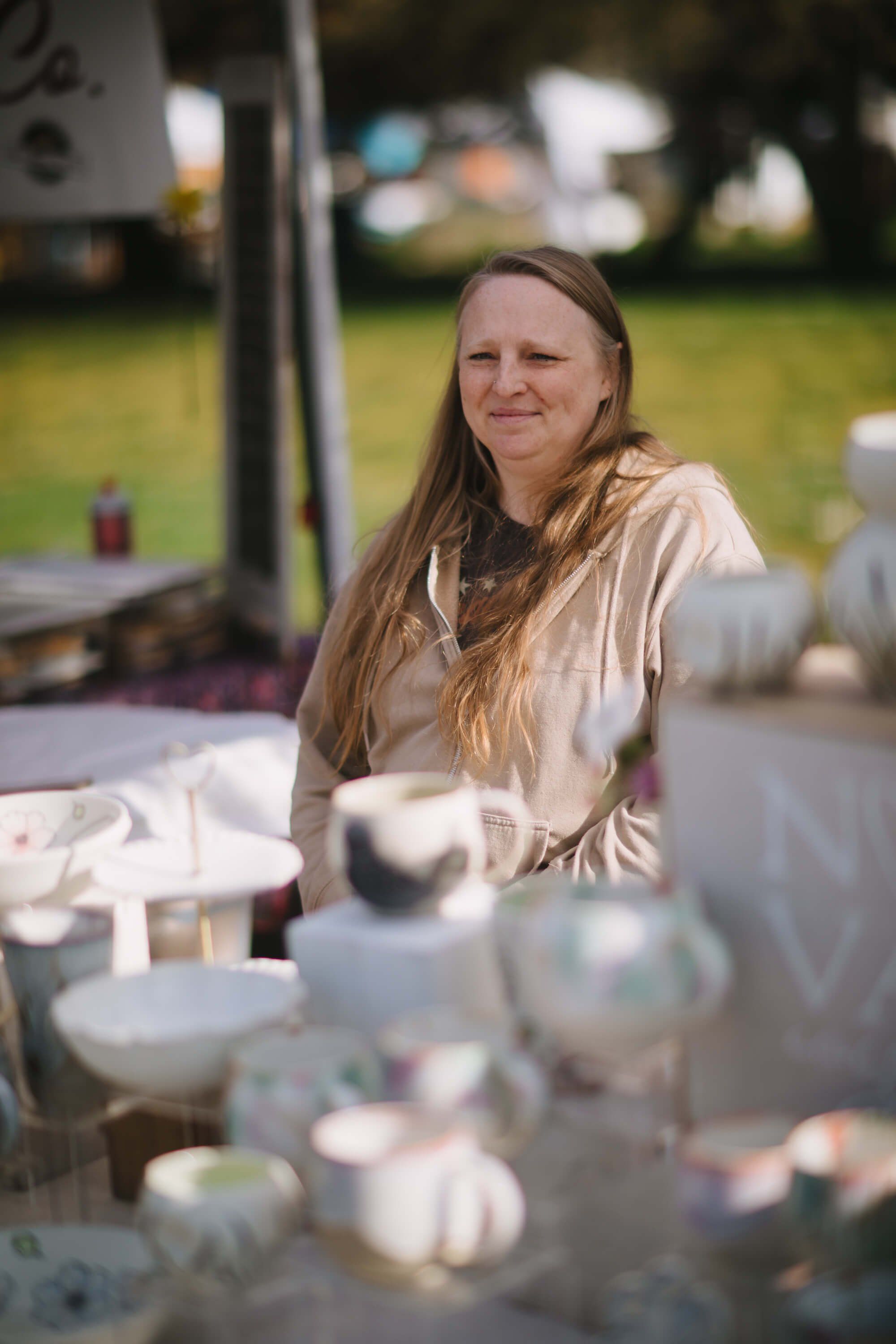 Woman with long hair at outdoor market table, soft afternoon light, green lawn and vendor tents in background