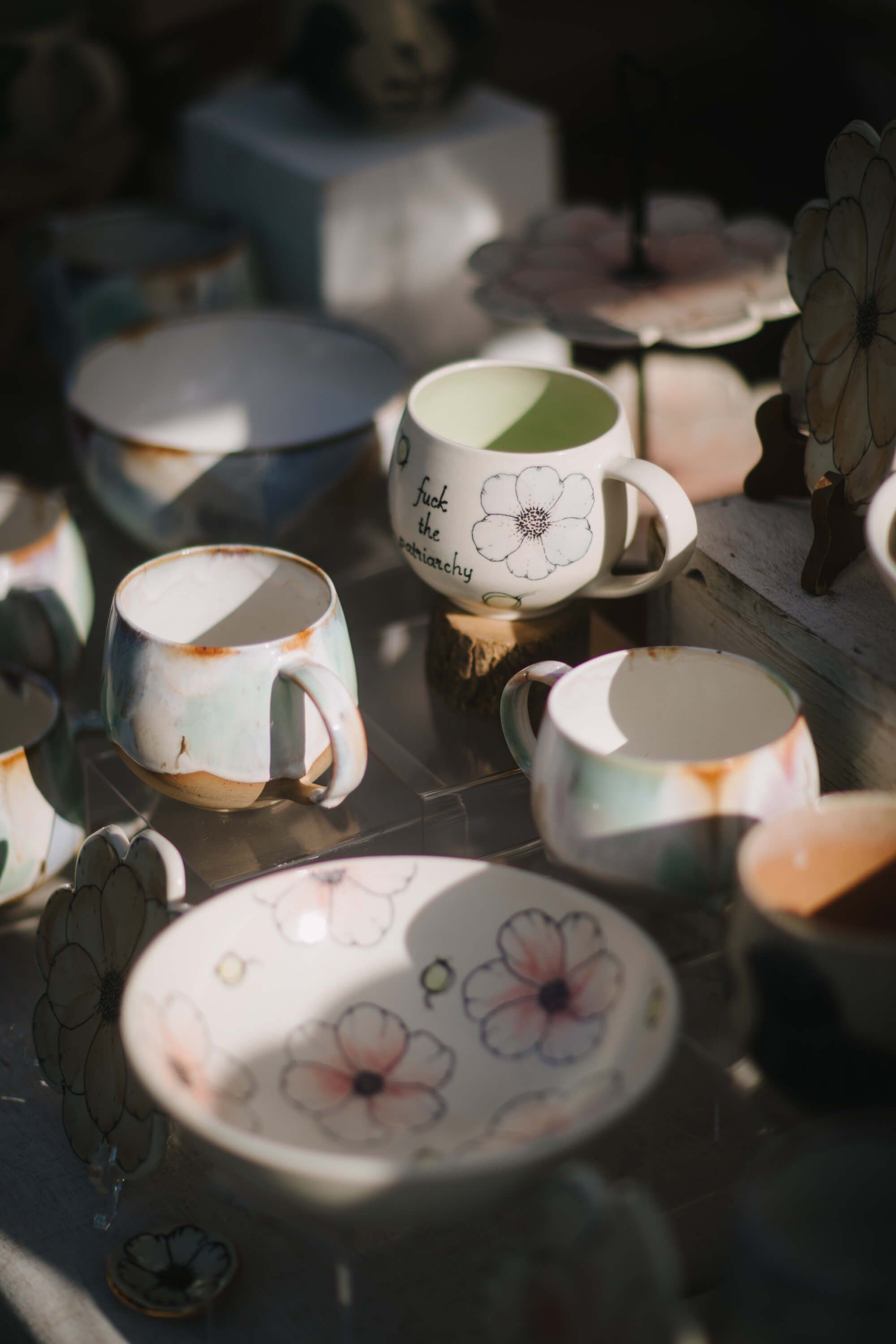 Handpainted ceramic mugs and plates with delicate floral designs arranged in dappled sunlight at an outdoor market