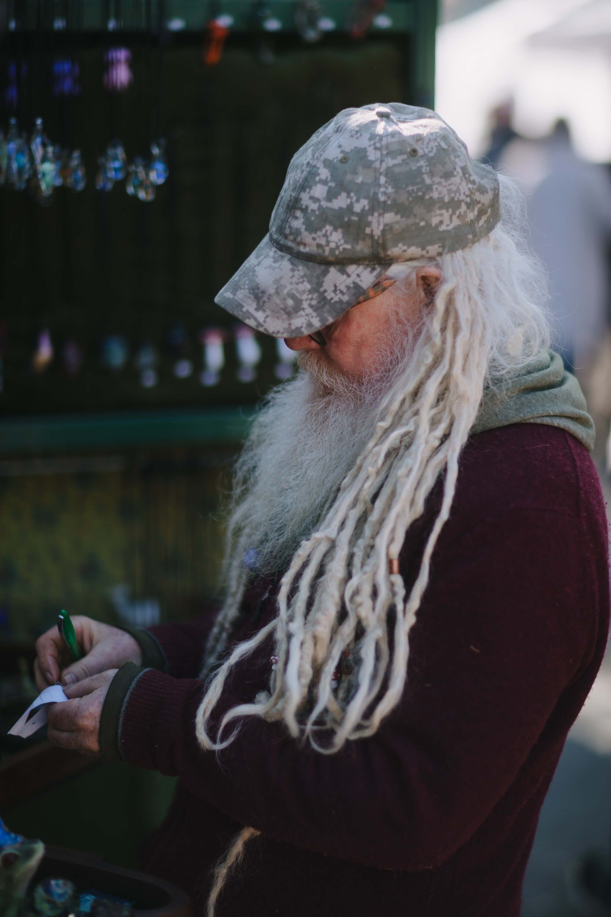Elderly man with long white dreadlocks and patterned cap examines something in his hands at an outdoor market, soft natural light
