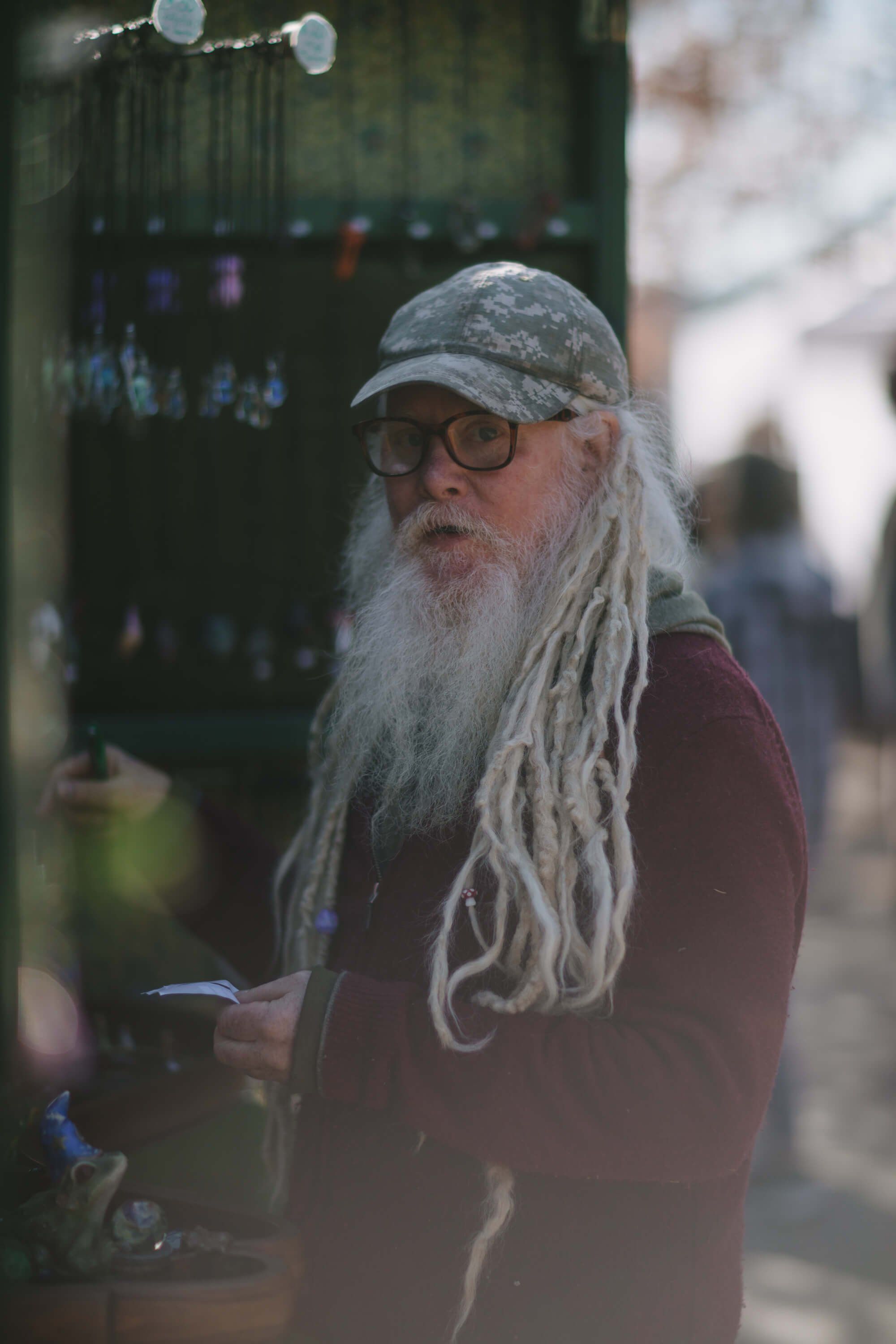 Bearded man with long gray dreadlocks and glasses at an outdoor market, wearing a burgundy shirt and cap