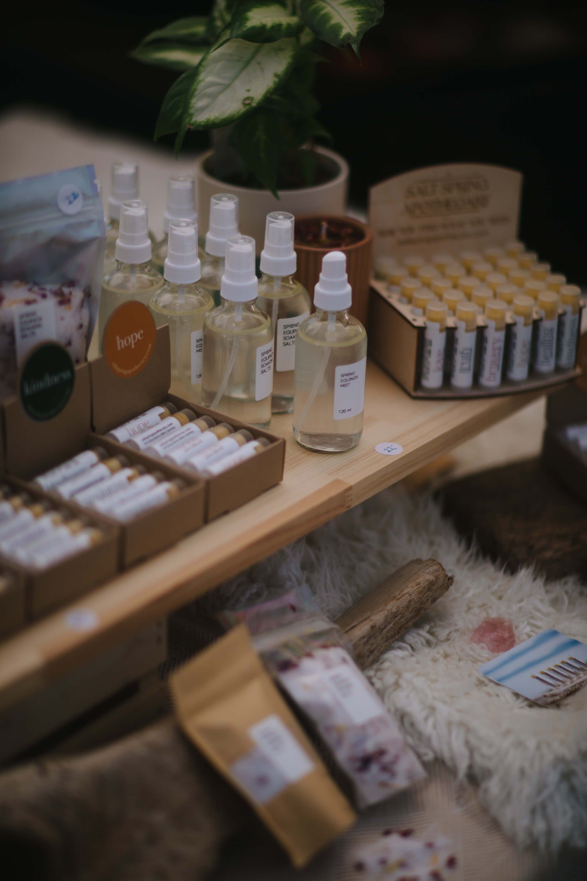 Artisan skincare products displayed on wooden shelves at market stall, featuring glass bottles, cardboard packaging, and small white product samples arranged…
