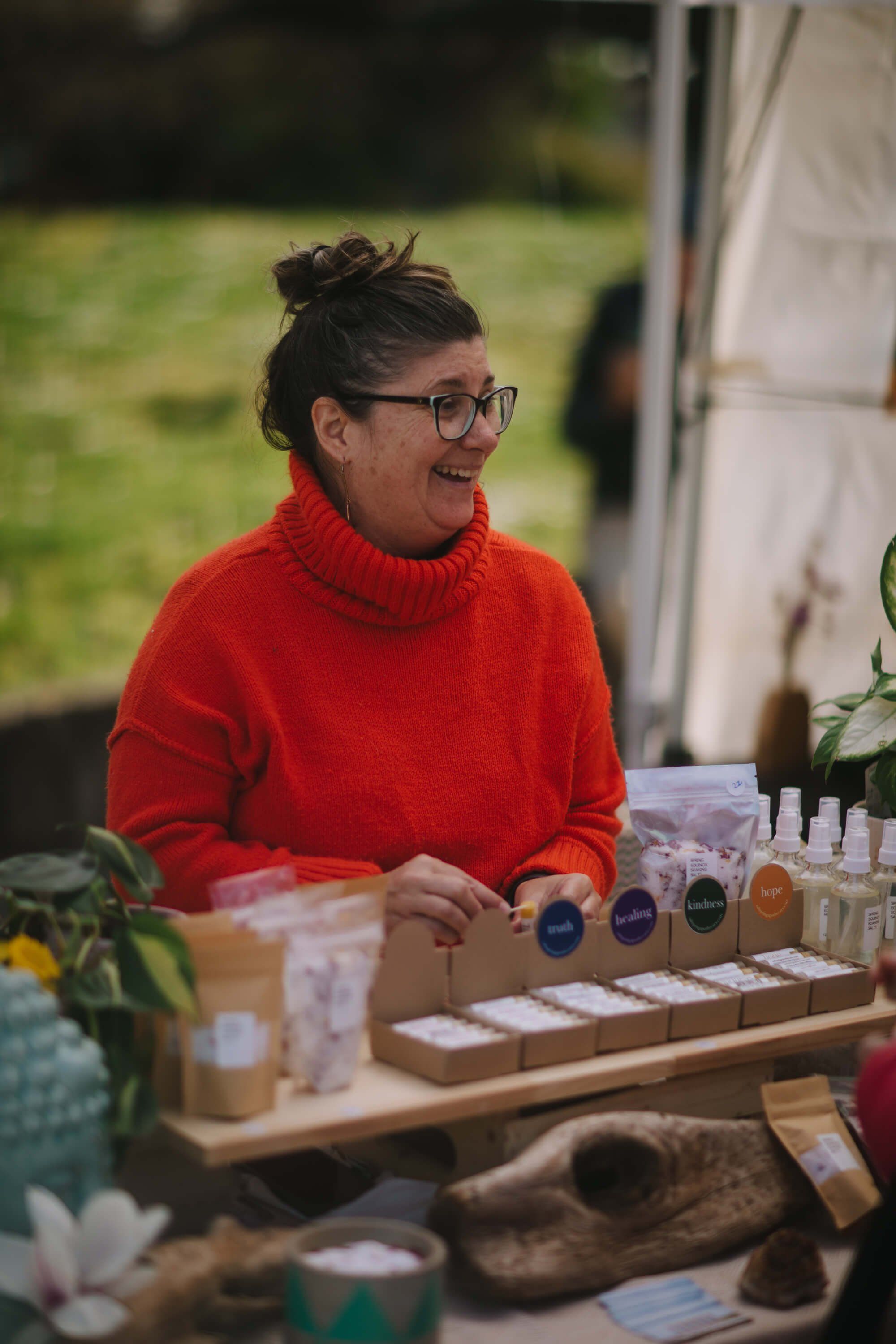 Woman in red turtleneck at market vendor table displaying glass bottles and natural products, smiling beside potted plants
