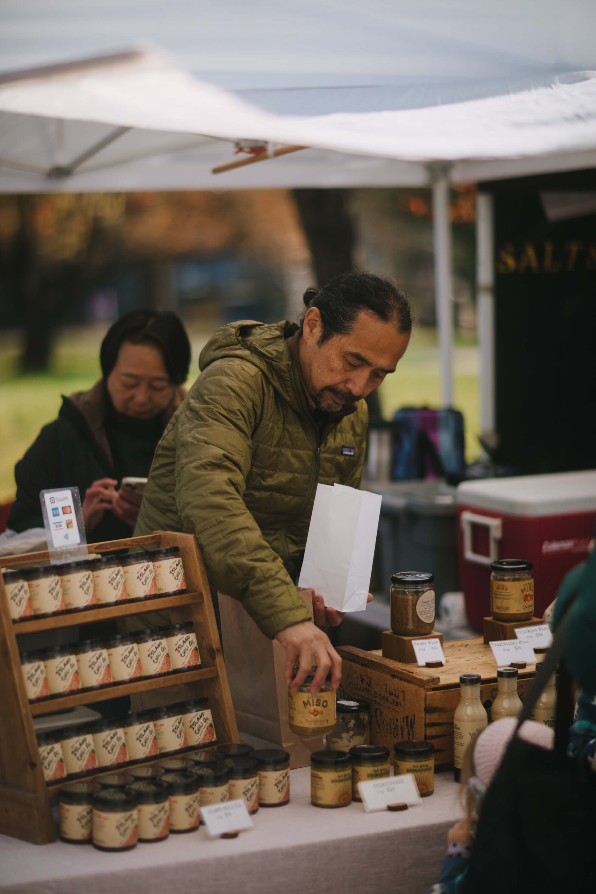 Vendor in olive jacket arranges jars at farmers market stall under white tent canopy