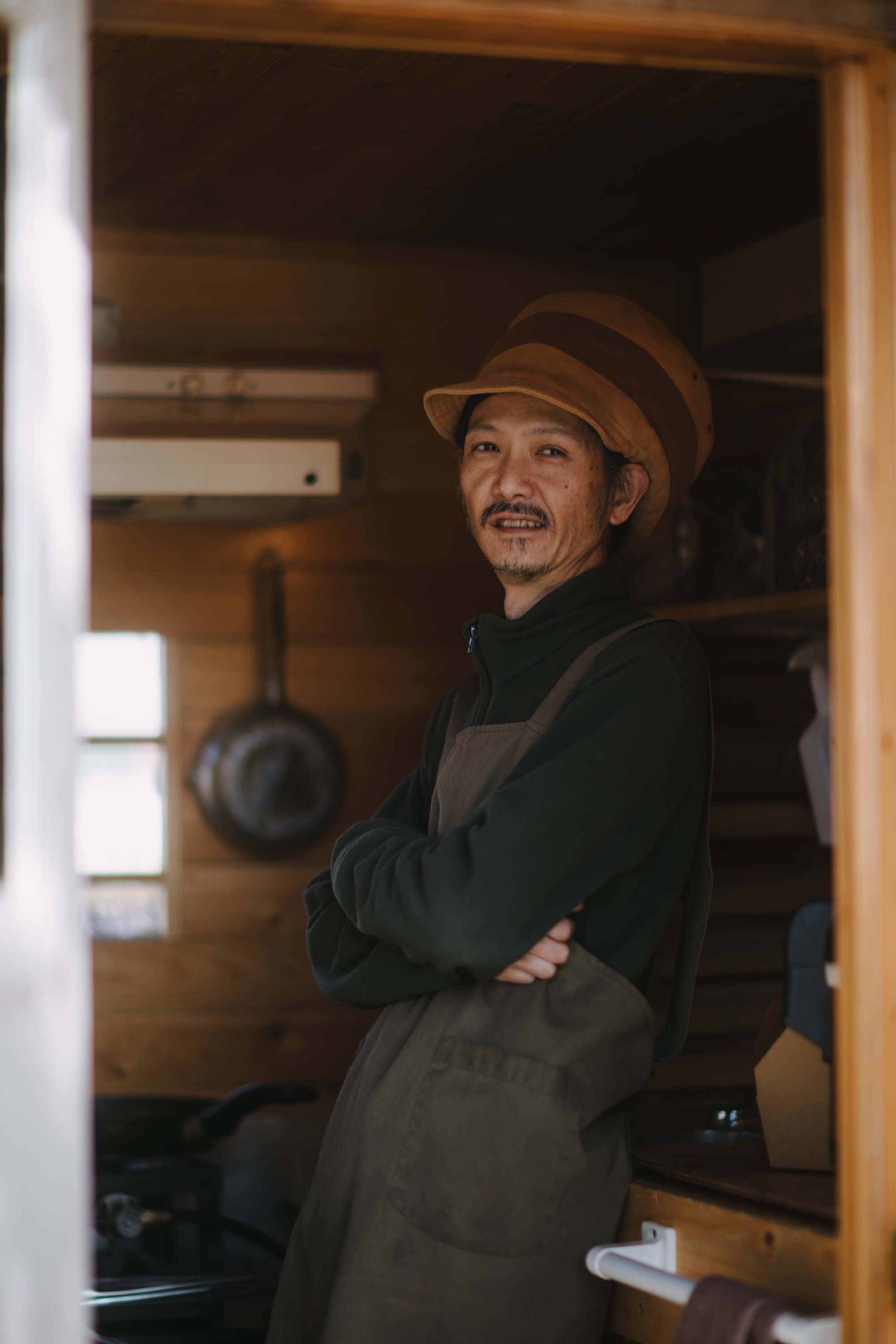 Man in striped hat and apron smiles warmly in a wooden kitchen doorway with cast iron cookware visible behind him