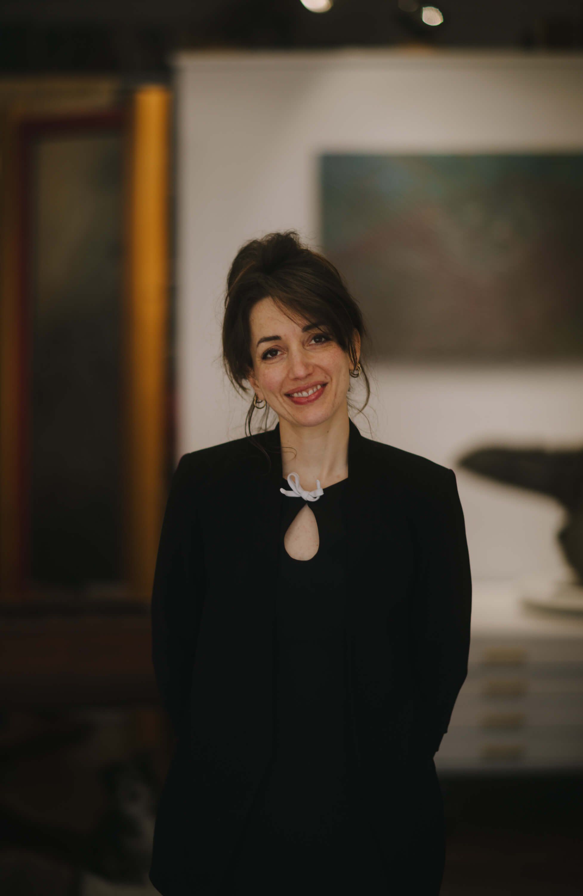 Woman in black blazer with red lipstick smiles at camera in softly lit indoor gallery space