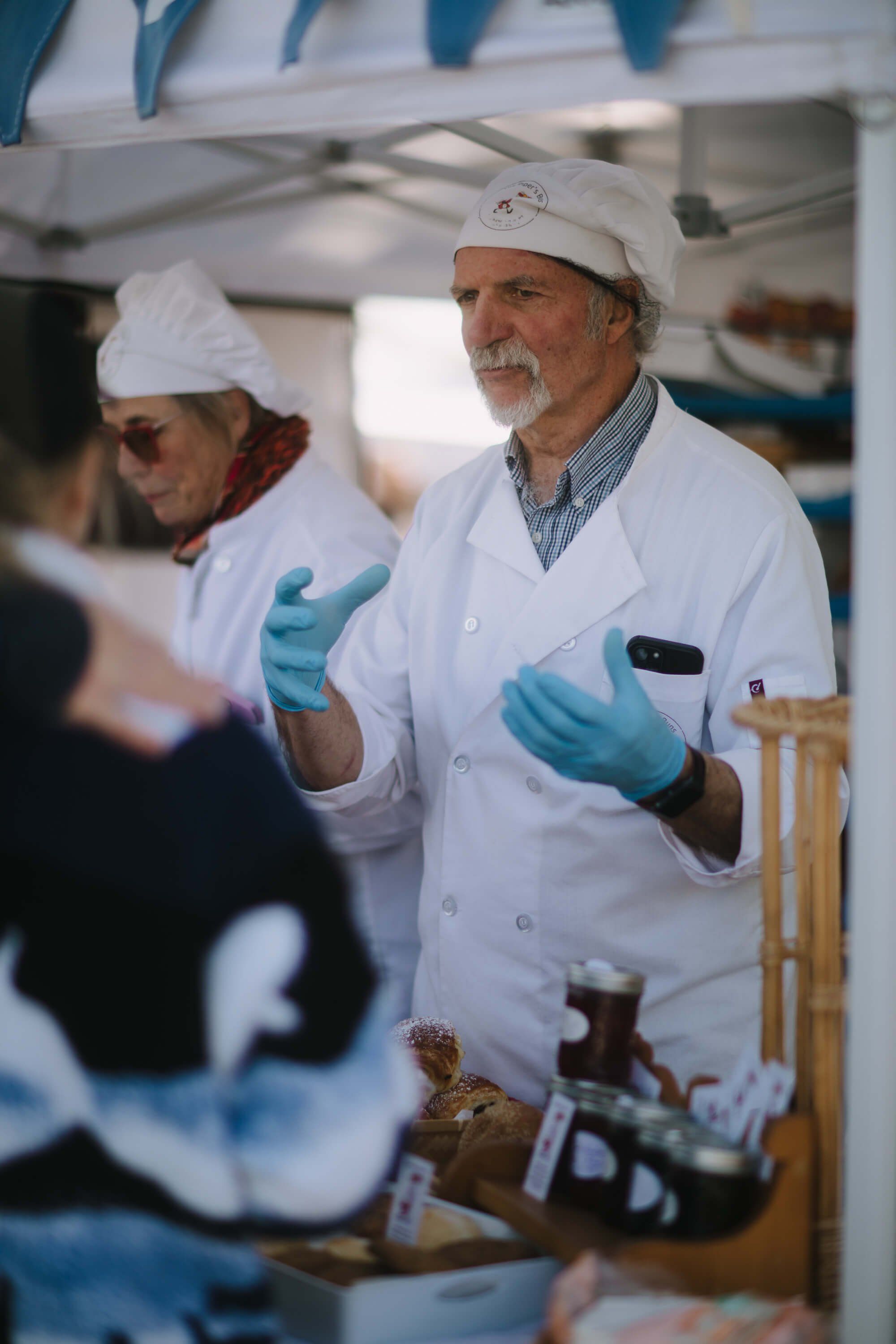 Man in white chef's coat and headwrap gestures while speaking at a market food preparation station, wearing blue gloves