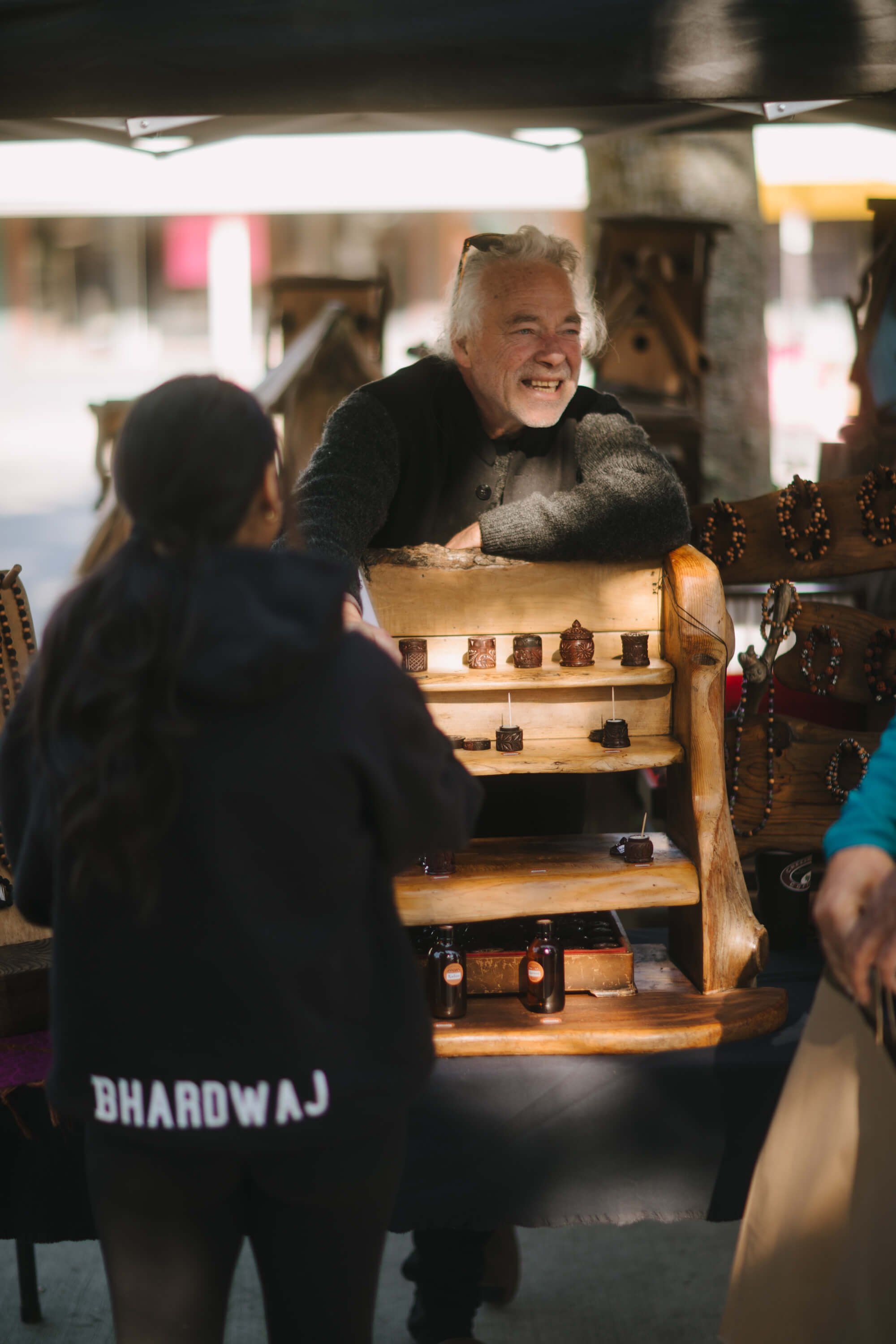 Vendor with silver hair smiles while showing jewelry display to customer at outdoor market stall
