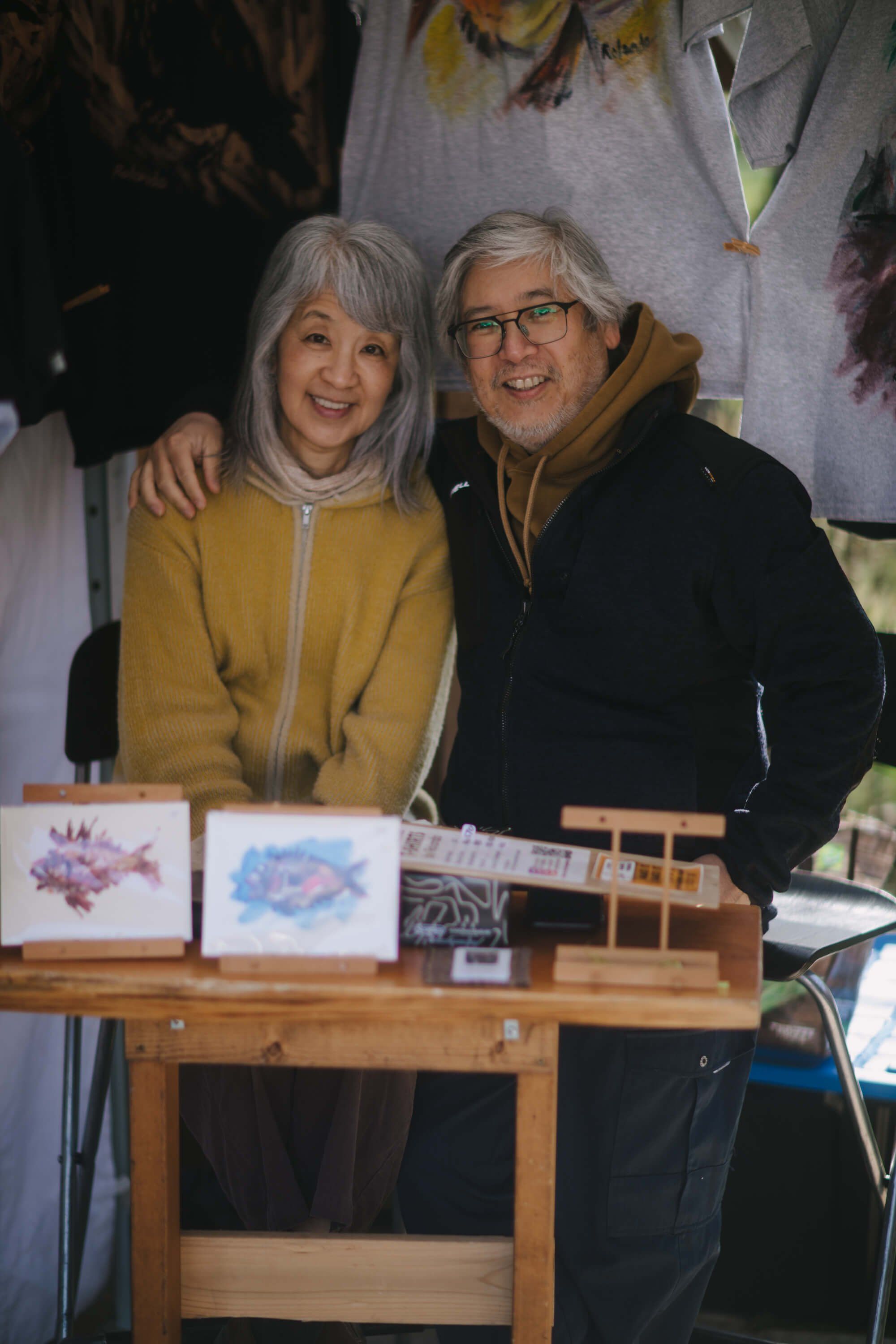 Two smiling vendors at a market booth displaying artwork, standing behind a wooden display table with framed pieces