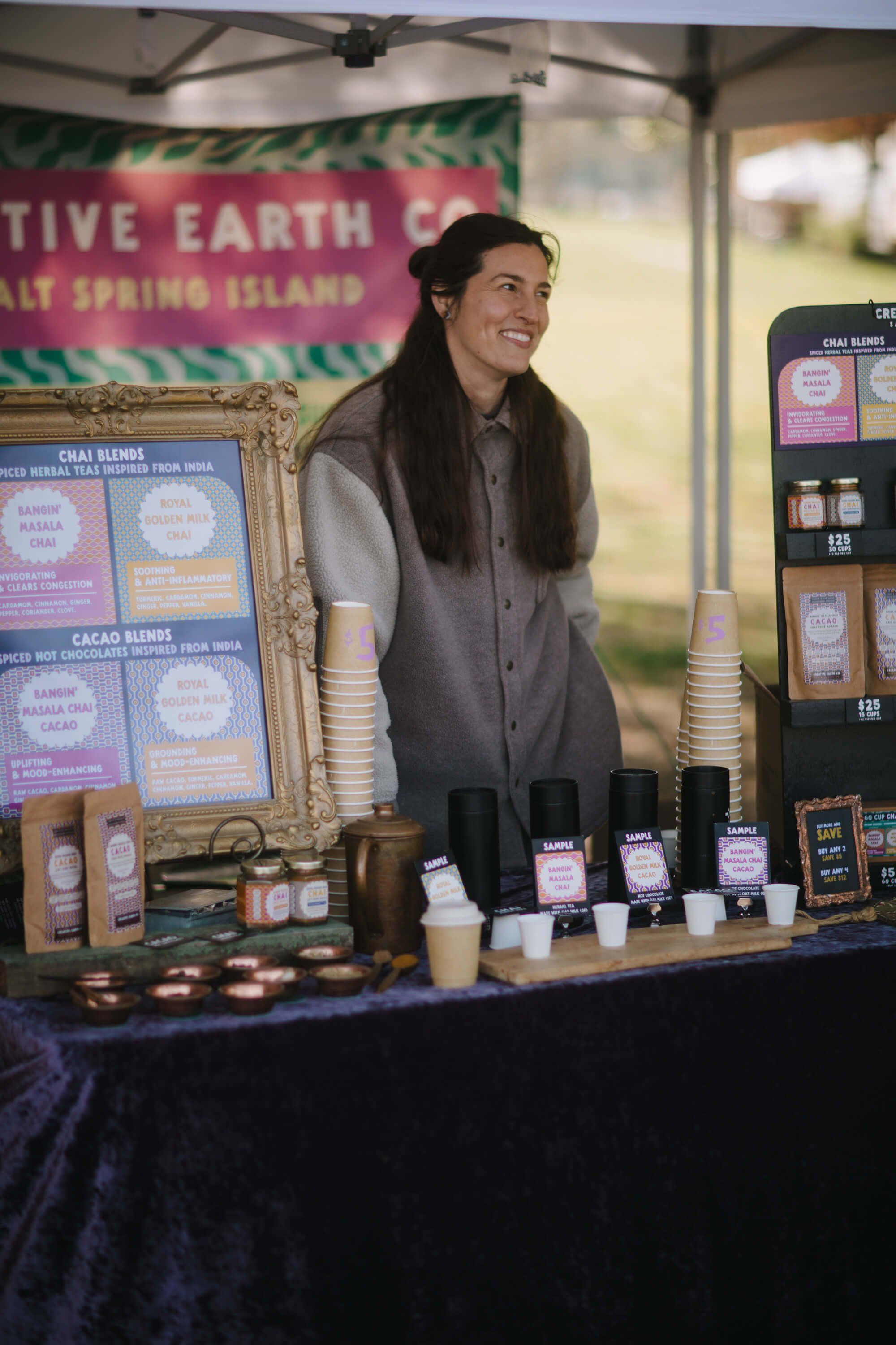 Vendor at market booth with product displays, smiling while tending to handmade goods under covered market tent