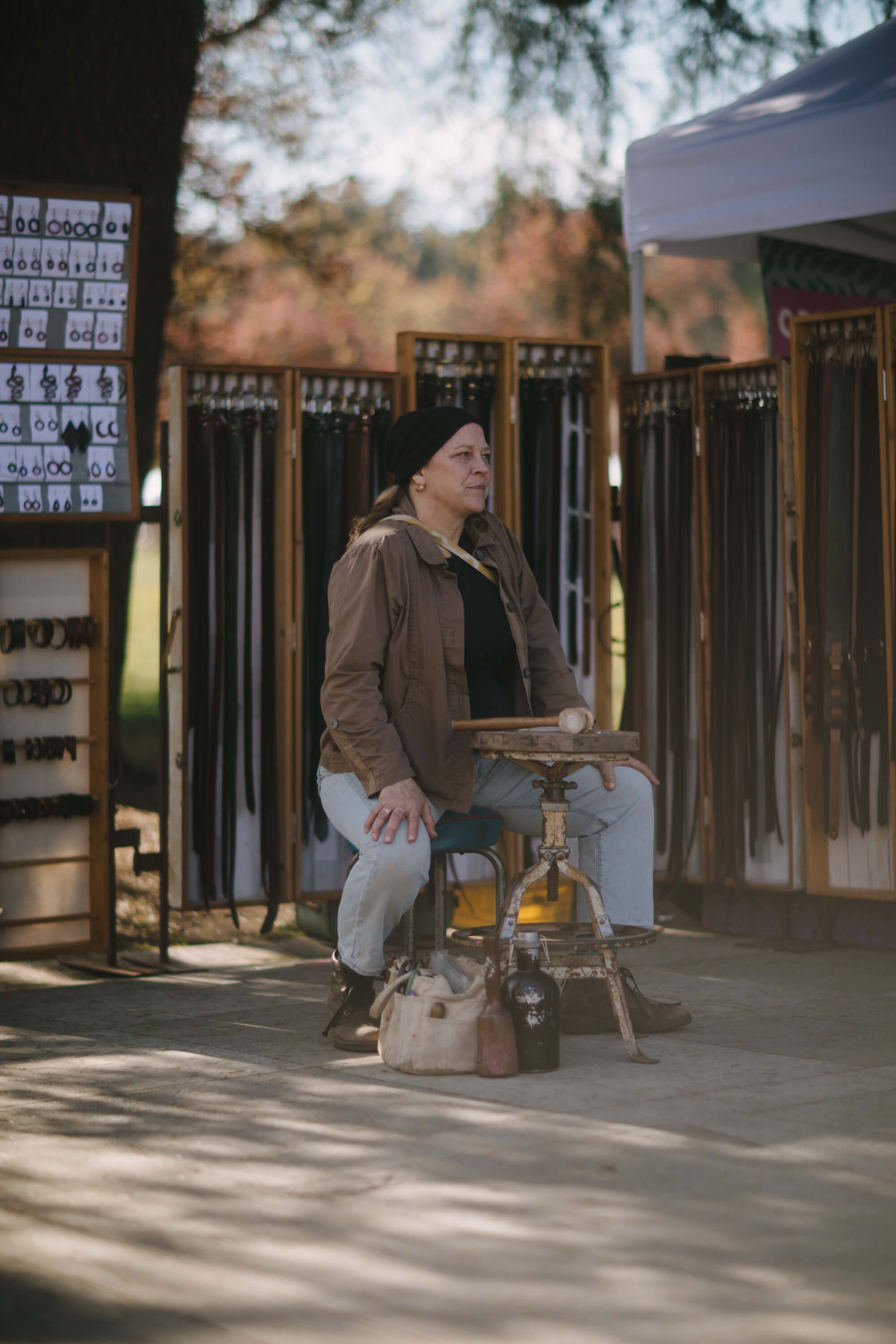 Person seated on wooden stool at outdoor market booth with fabric displays, dappled sunlight filtering through trees