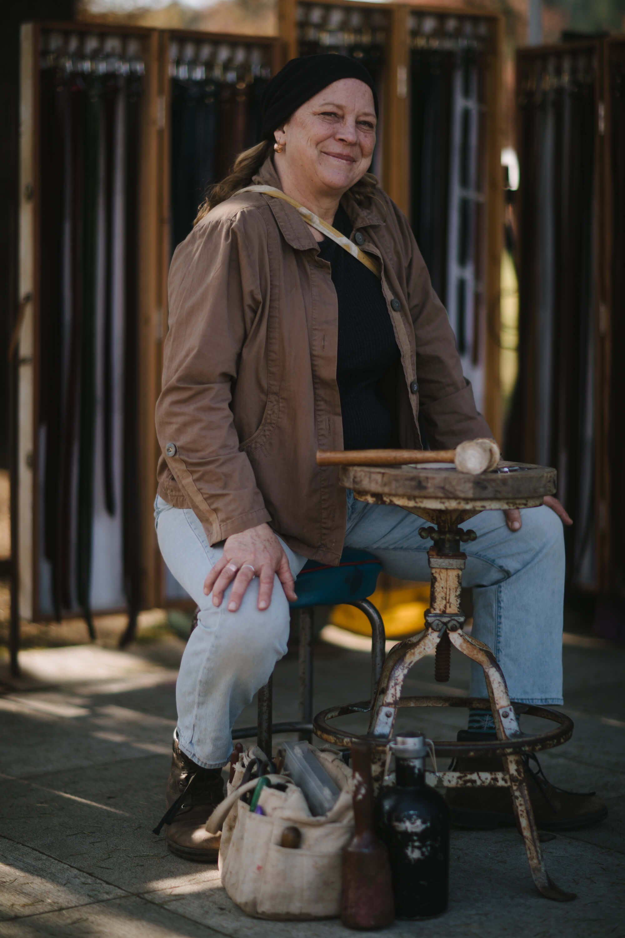 Vendor smiles while sitting at outdoor market stall with drum among displayed goods, warm sunlight casting shadows behind wooden structures