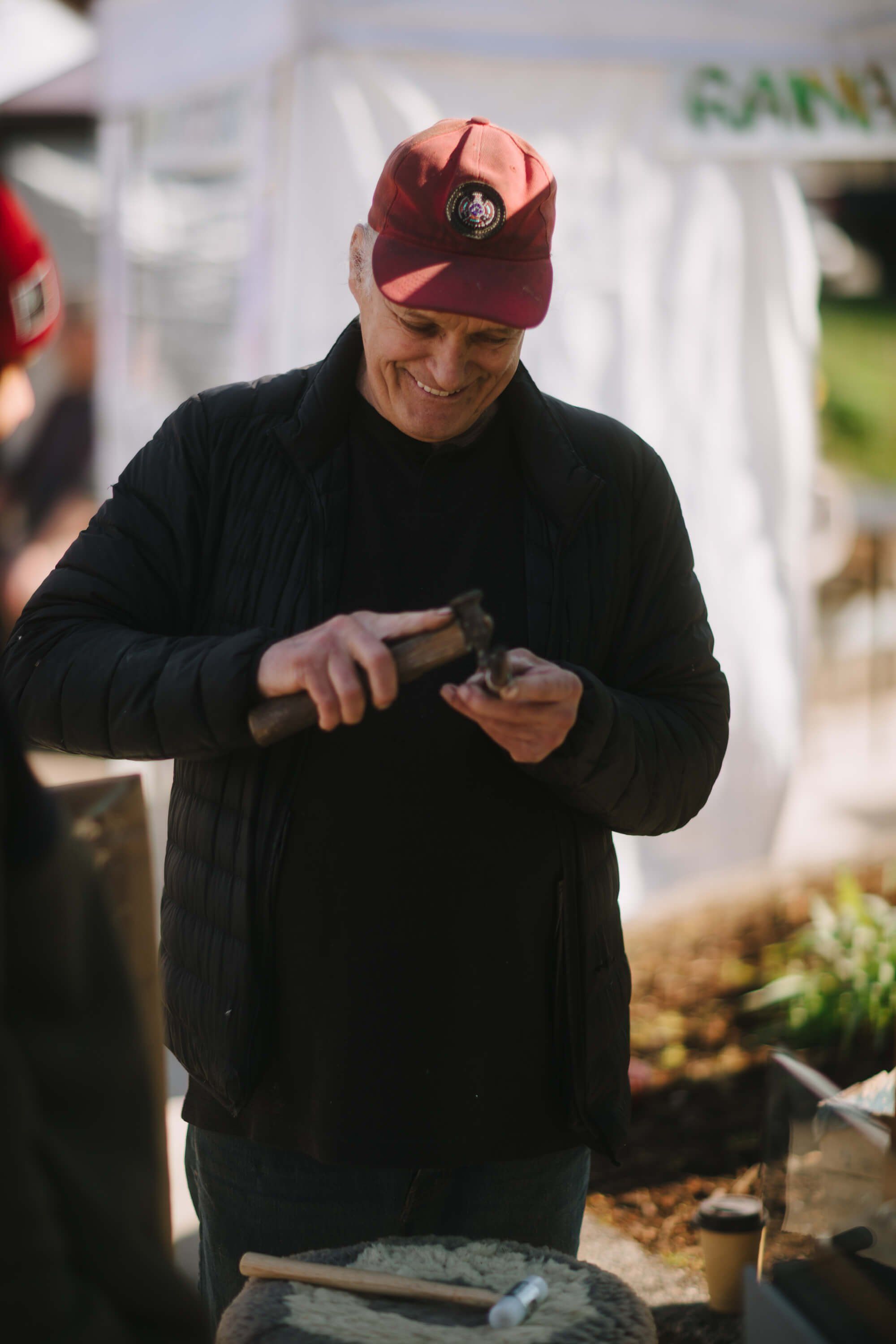 Man in maroon cap and black jacket examines produce at outdoor market stall in natural daylight