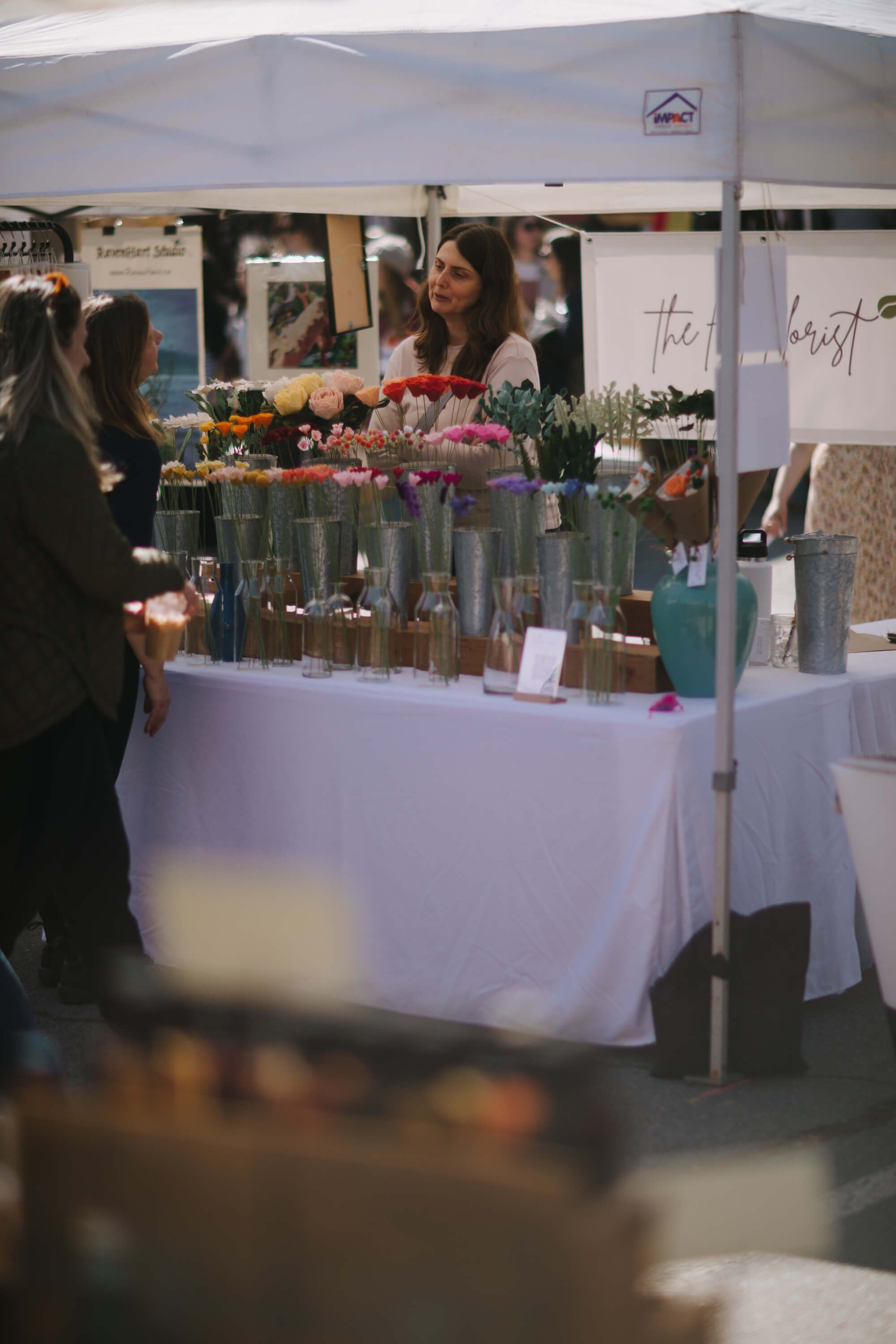 Vendor at outdoor market tent displays glass bottles and floral arrangements on white-draped table while attending customer