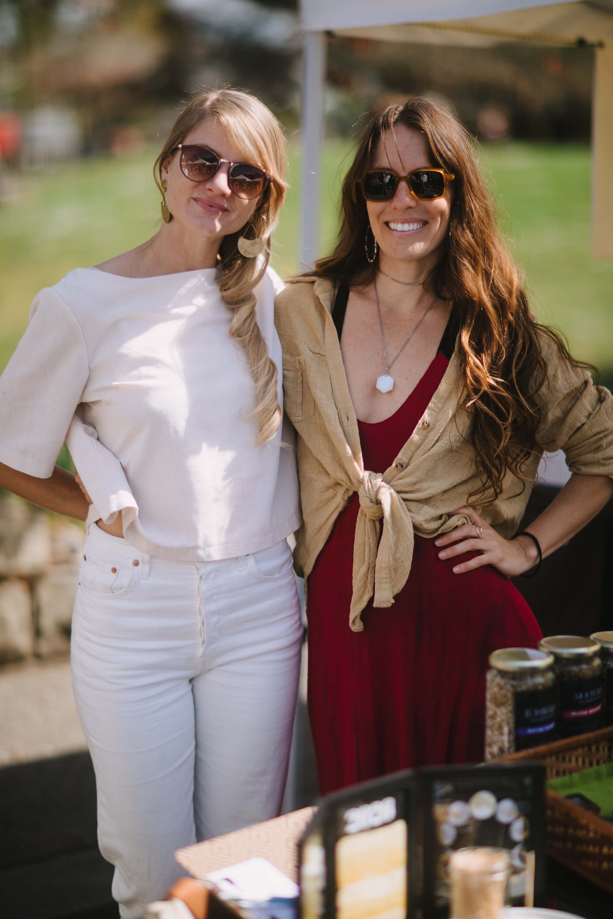 Two women in sunglasses smile at the market in bright afternoon sunlight, one wearing white and the other a burgundy dress with tan cardigan