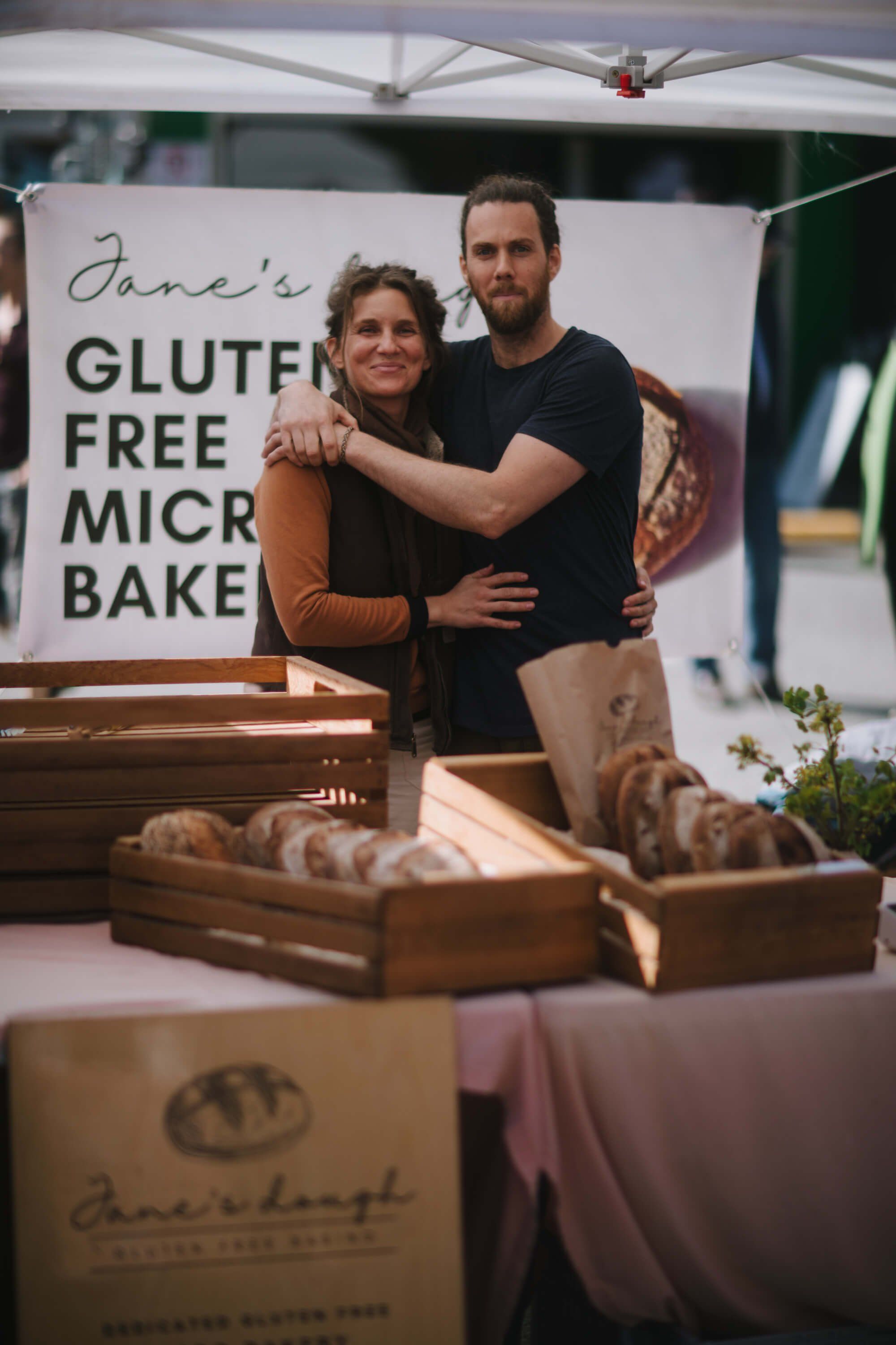 Vendors at farmers market stand behind gluten-free micro bakery display with fresh bread, warm natural light filtering through market tent
