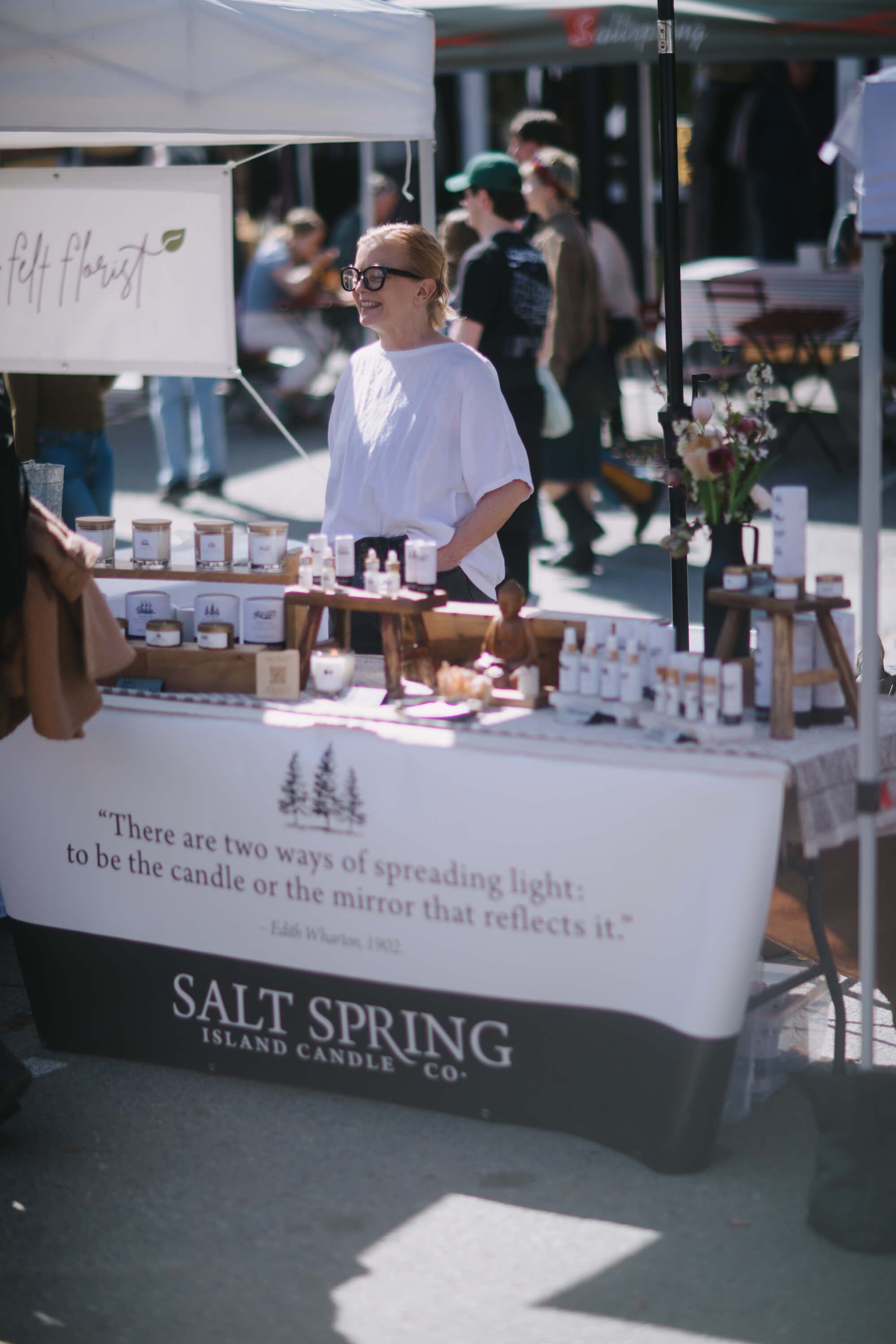 Vendor at sunny market booth displaying Salt Spring Island candles, with handwritten signage and product displays on white table