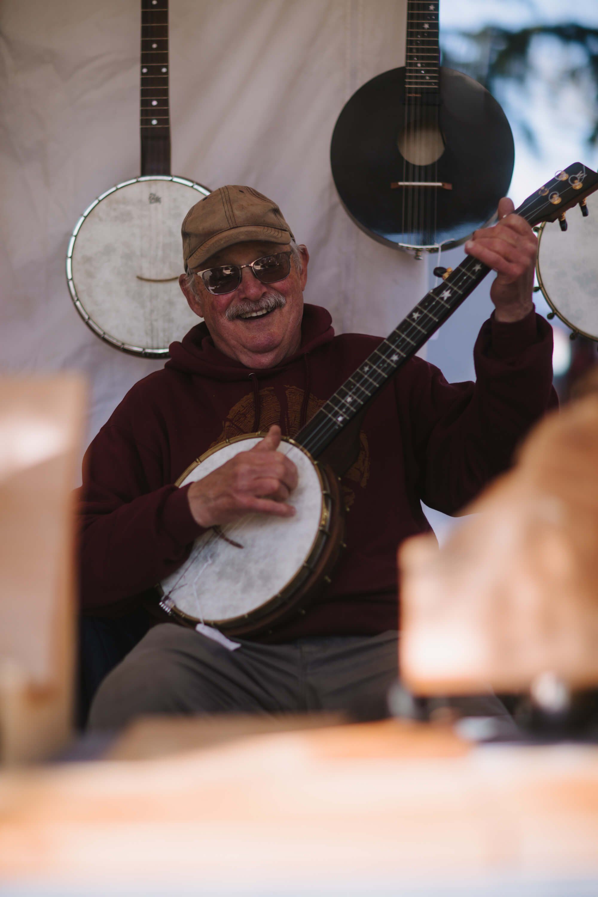 Banjo player wearing brown cap and glasses smiles while holding instrument, with acoustic guitars visible in soft-focus background at market stall