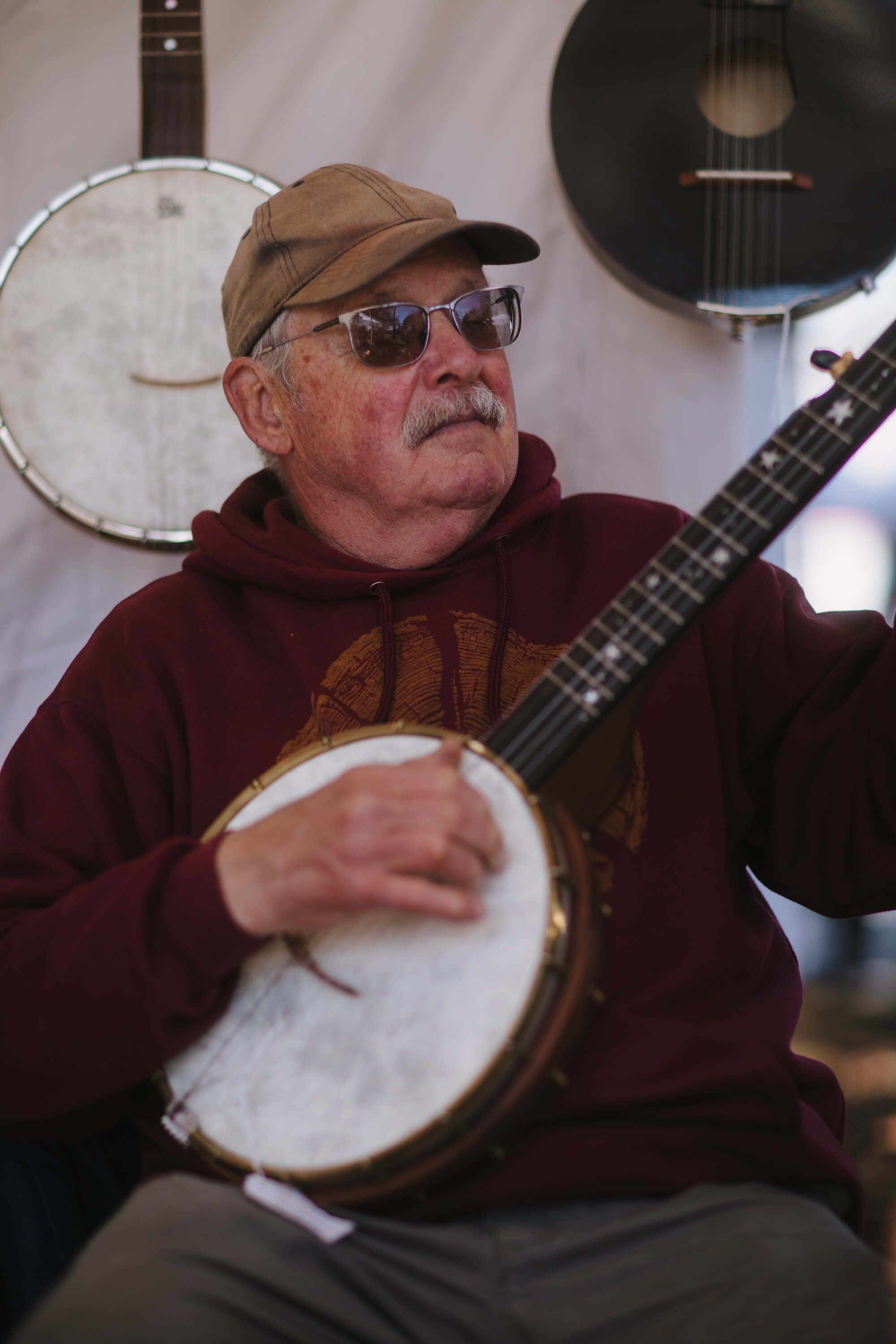 Musician wearing glasses and brown cap plays banjo at Saturday Market, with acoustic guitars visible in soft-focused background