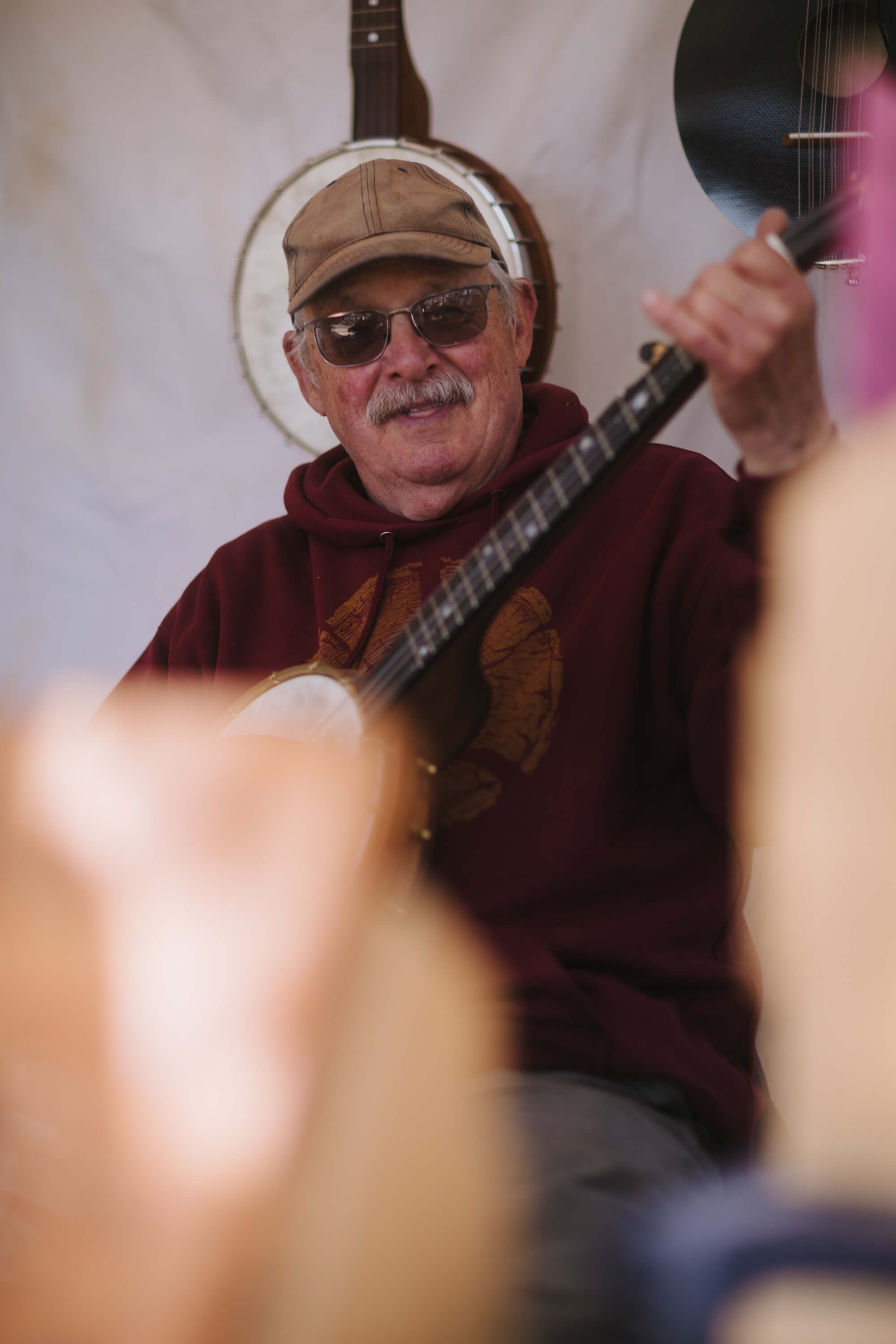Musician wearing sunglasses and cap plays acoustic guitar at Salt Spring Island Saturday Market, warm sunlight creating soft bokeh in foreground