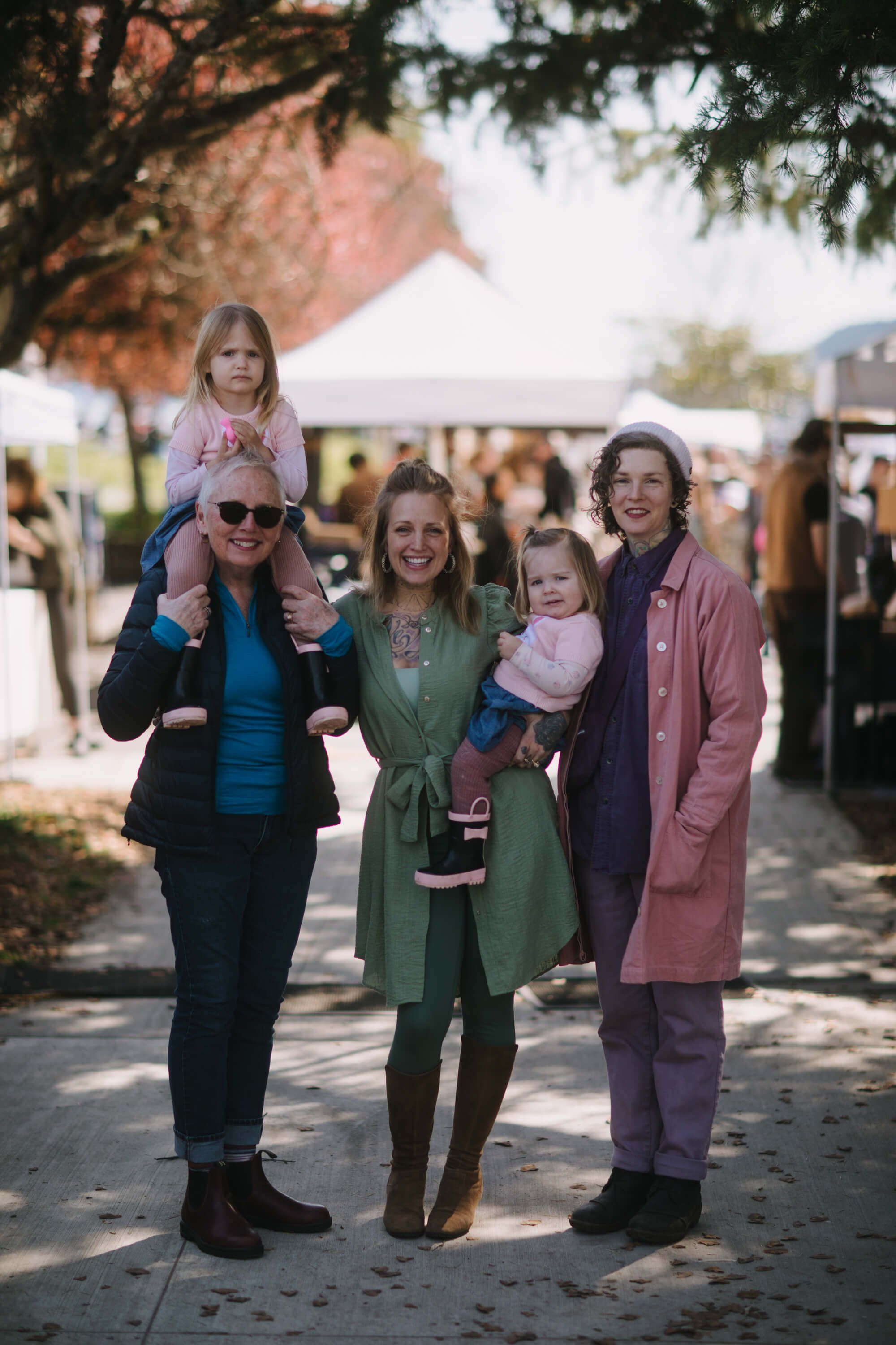 Family of five smiling under a tree at a sunny outdoor market, with tents and crowds visible in the warm background