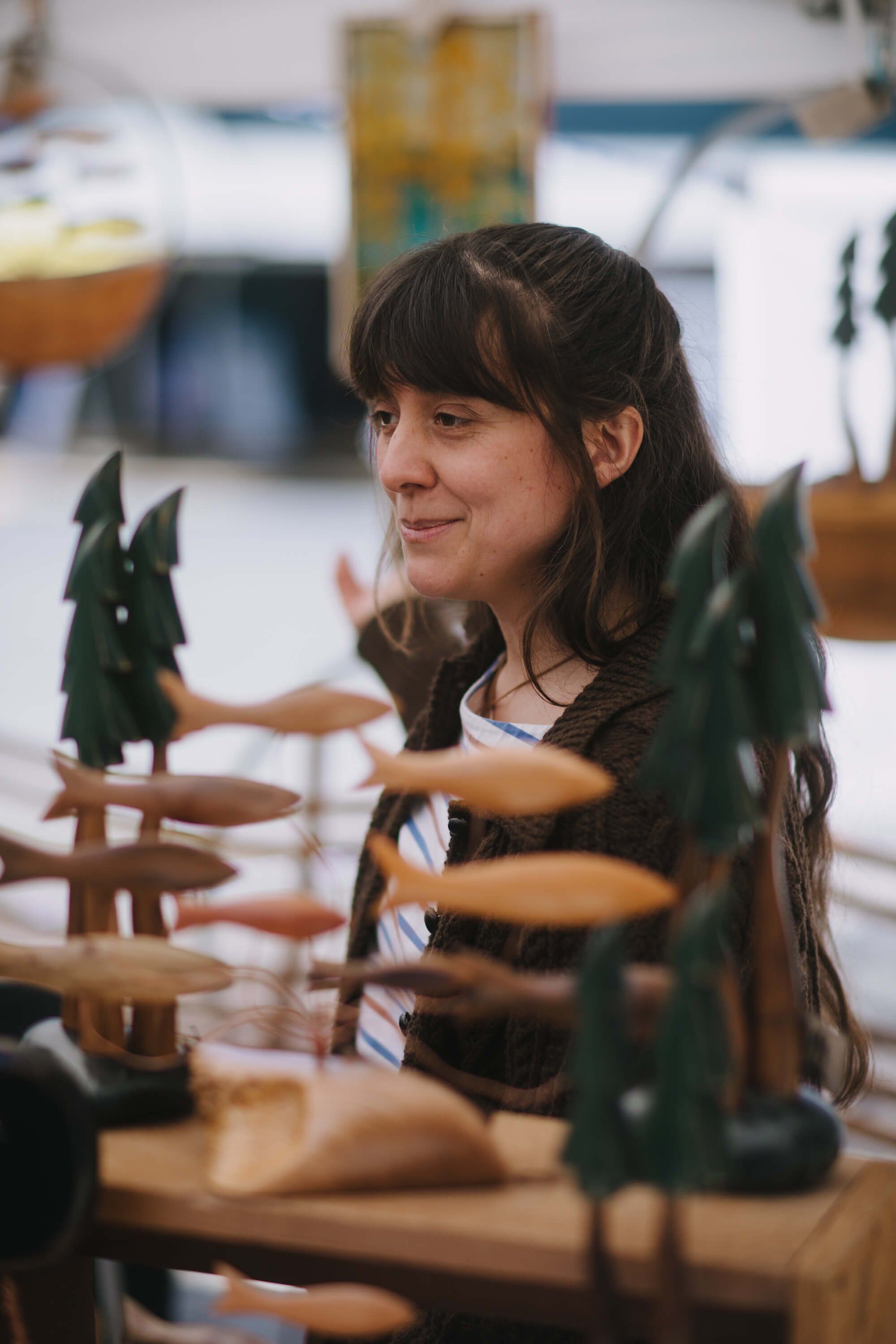 Woman with dark hair and bangs smiles among wooden carved figures at a market stall