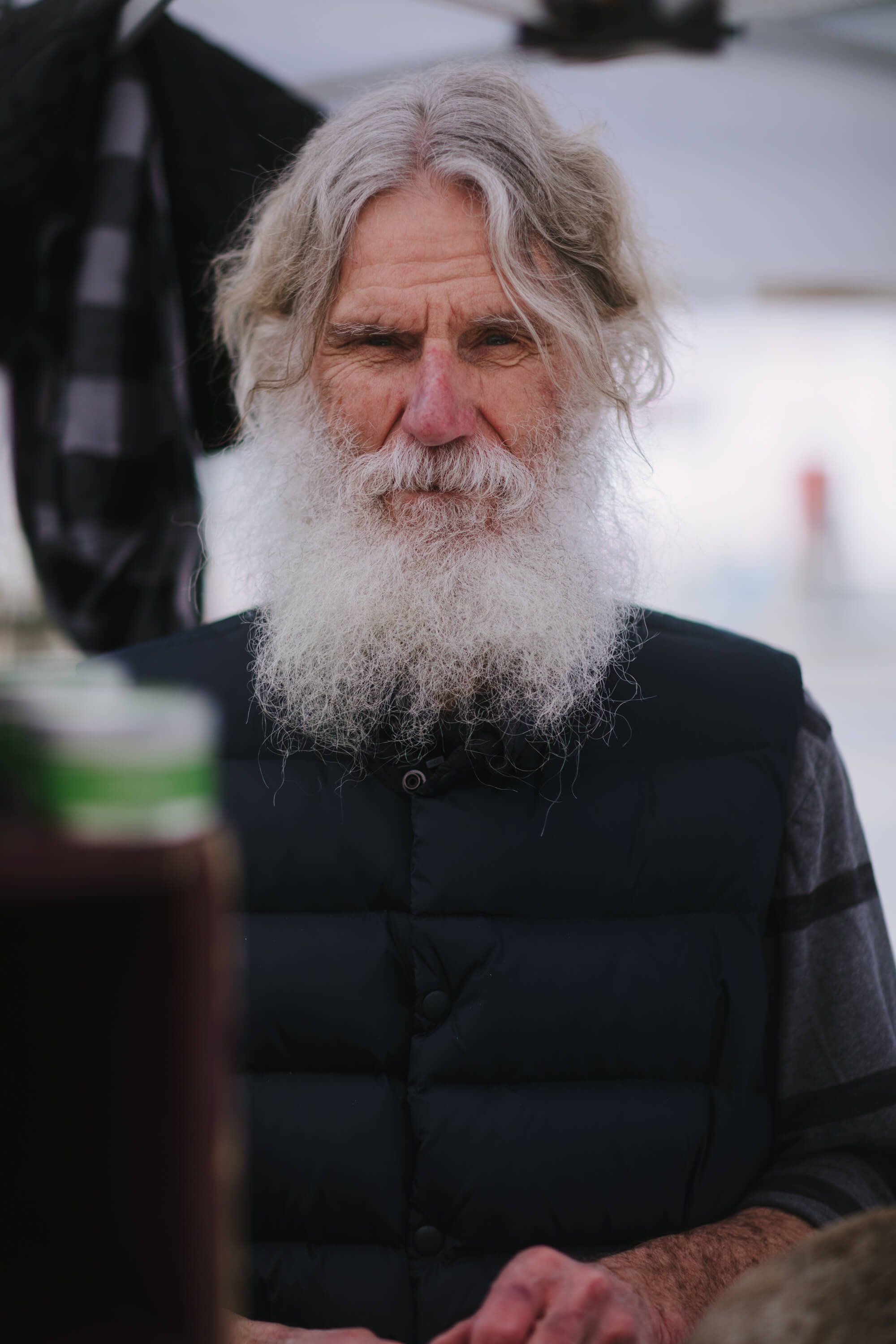 Older man with long white beard and grey hair wearing a dark vest, photographed outdoors in natural light at the Saturday Market