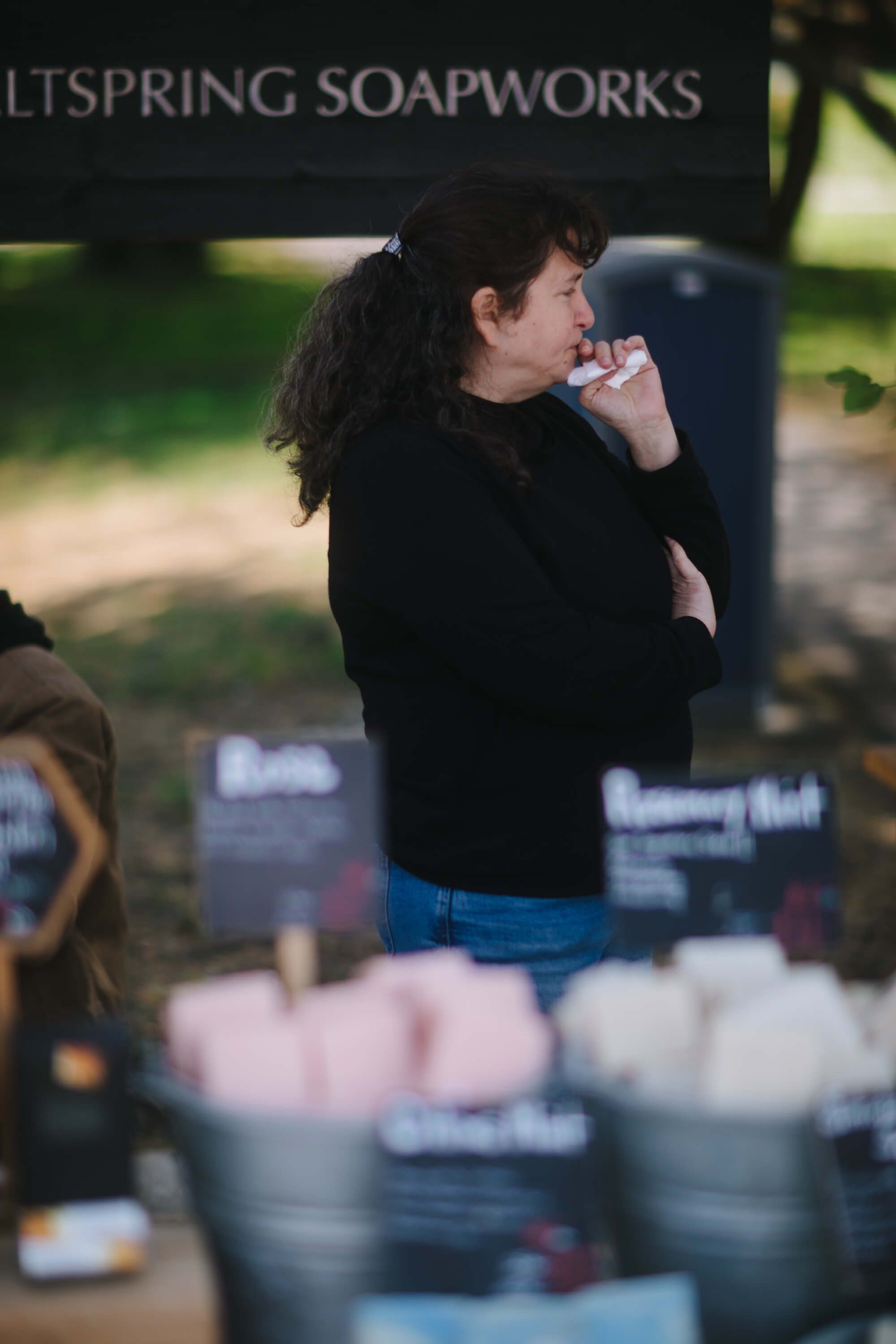 Person in black jacket sampling soap at Offspring Soapworks market stall, surrounded by colorful product displays