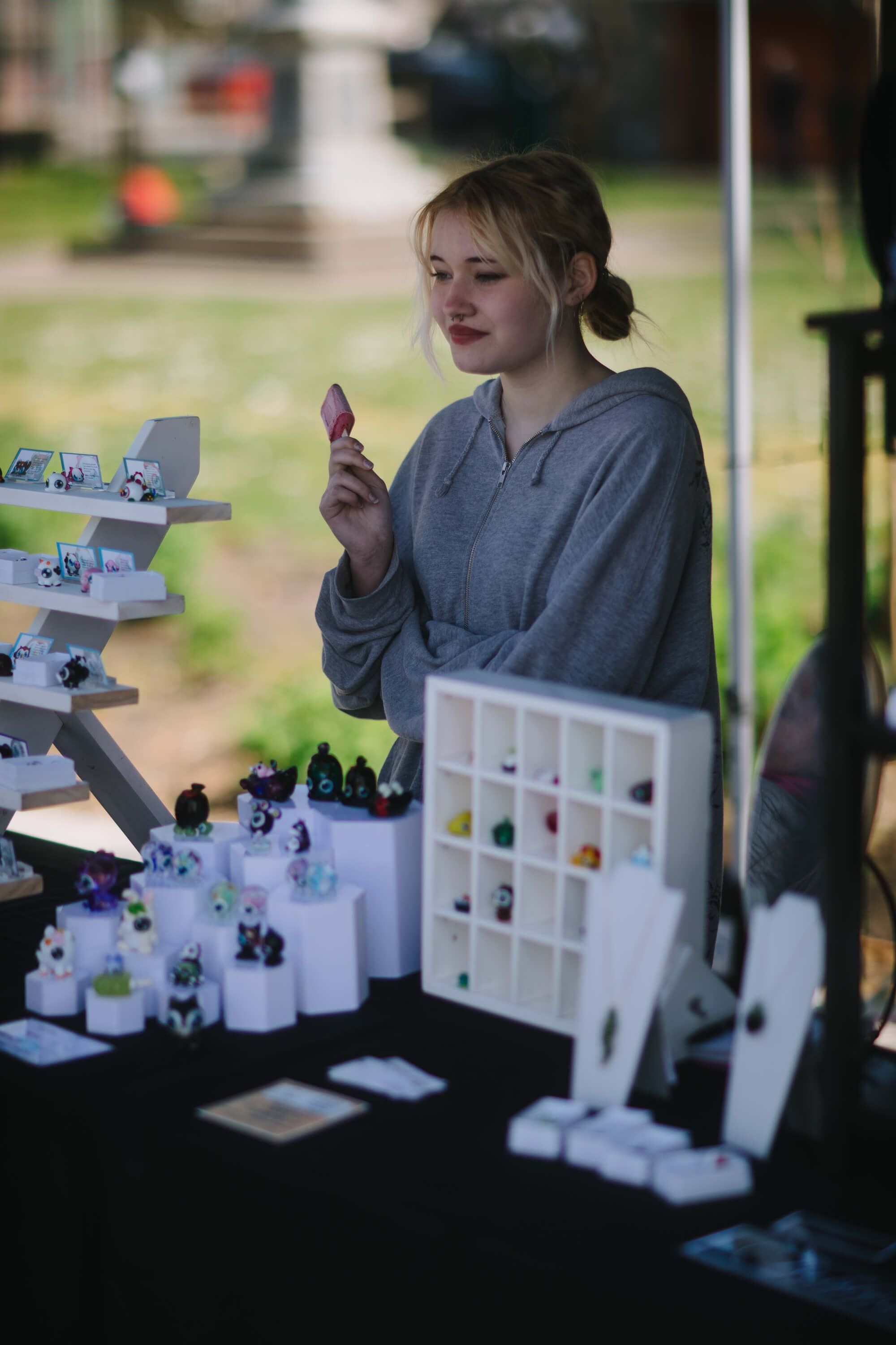 Young woman at outdoor market booth displaying handmade ceramics and small goods in natural daylight
