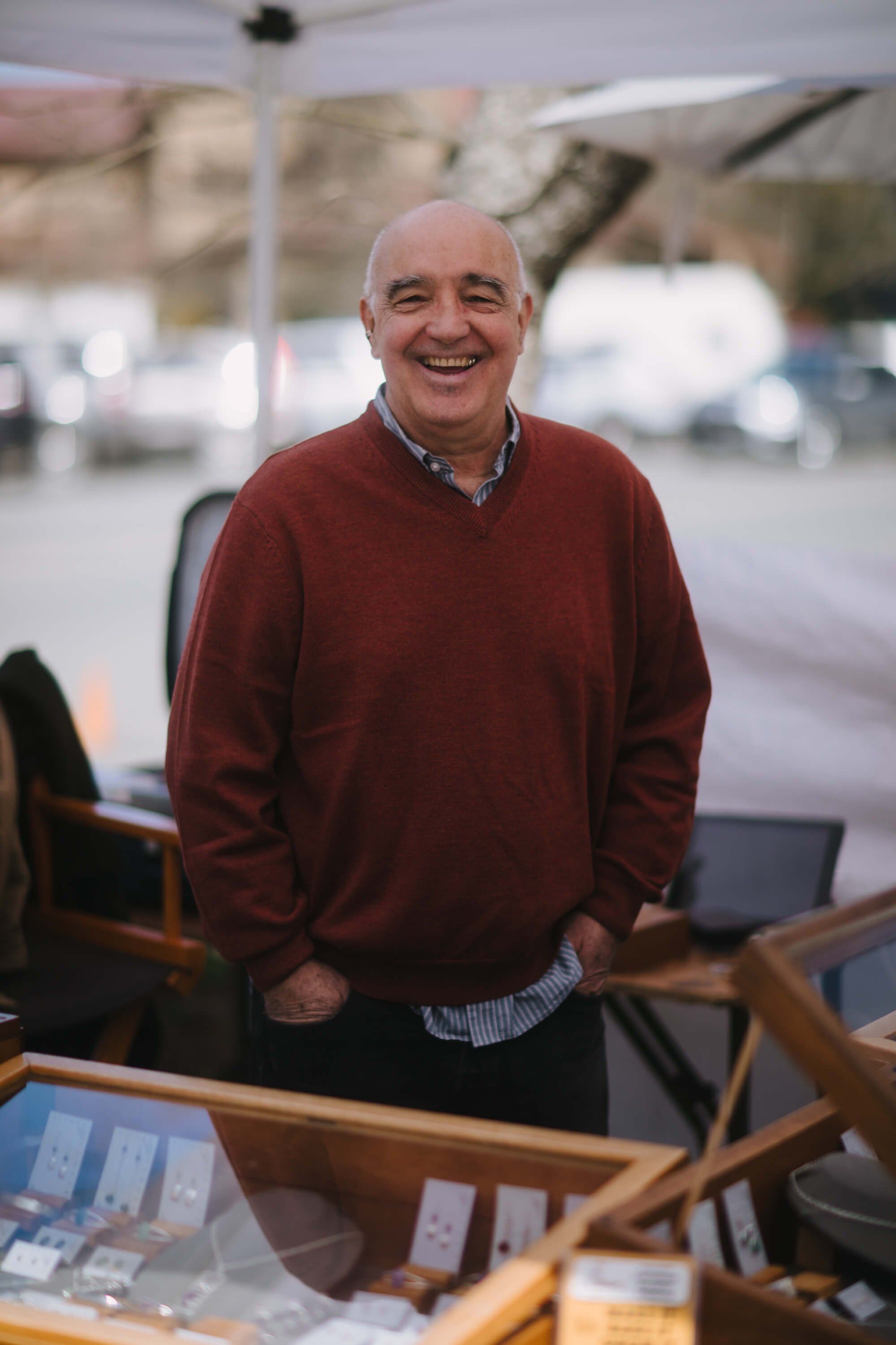 Smiling man in burgundy sweater stands at market stall with wooden display and product samples on table