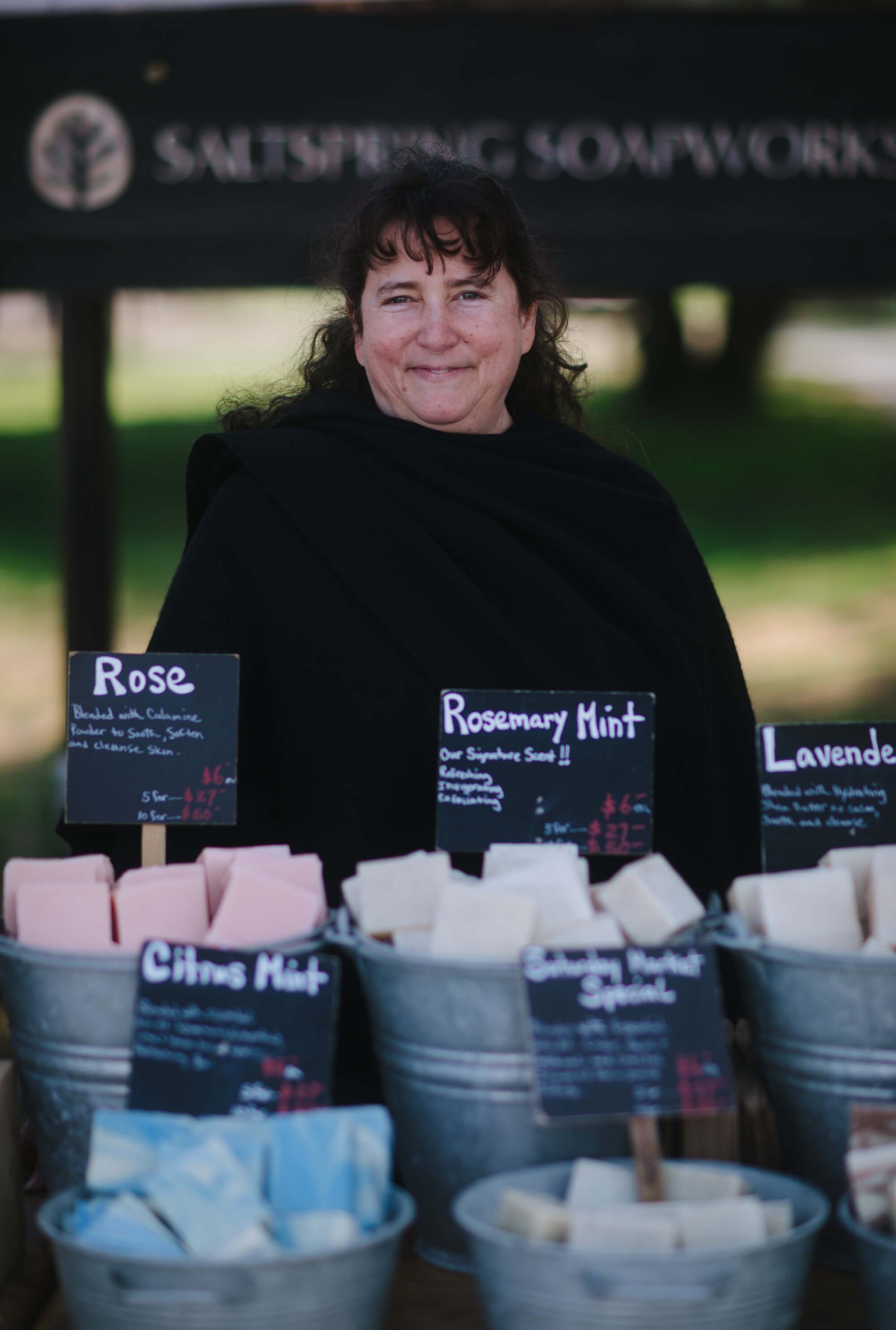 Woman smiling at craft market stall displaying handmade candles in various scents and pastel colors