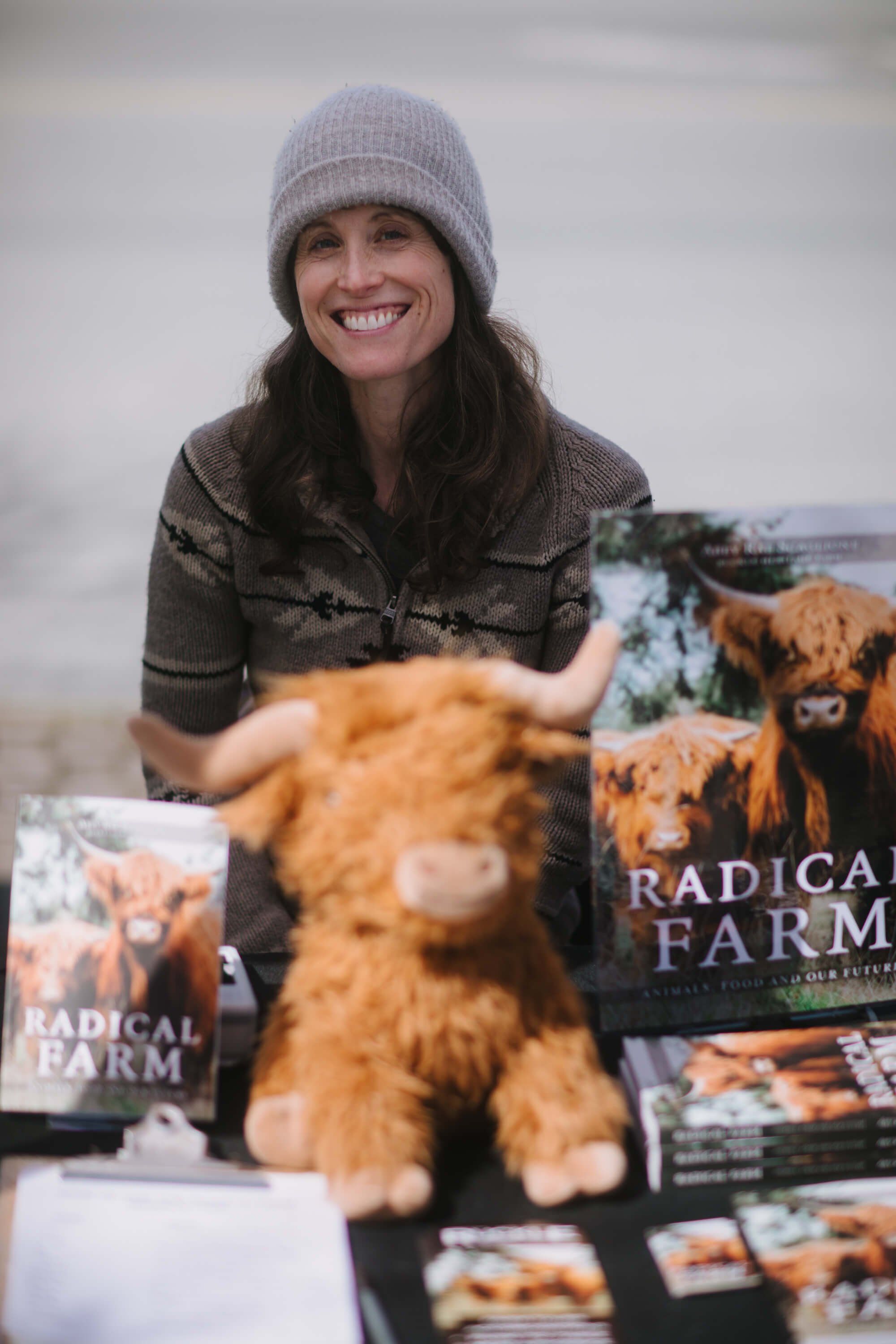 Smiling woman in gray beanie holds Highland cattle plush toy at market table displaying "Radical Farm" books