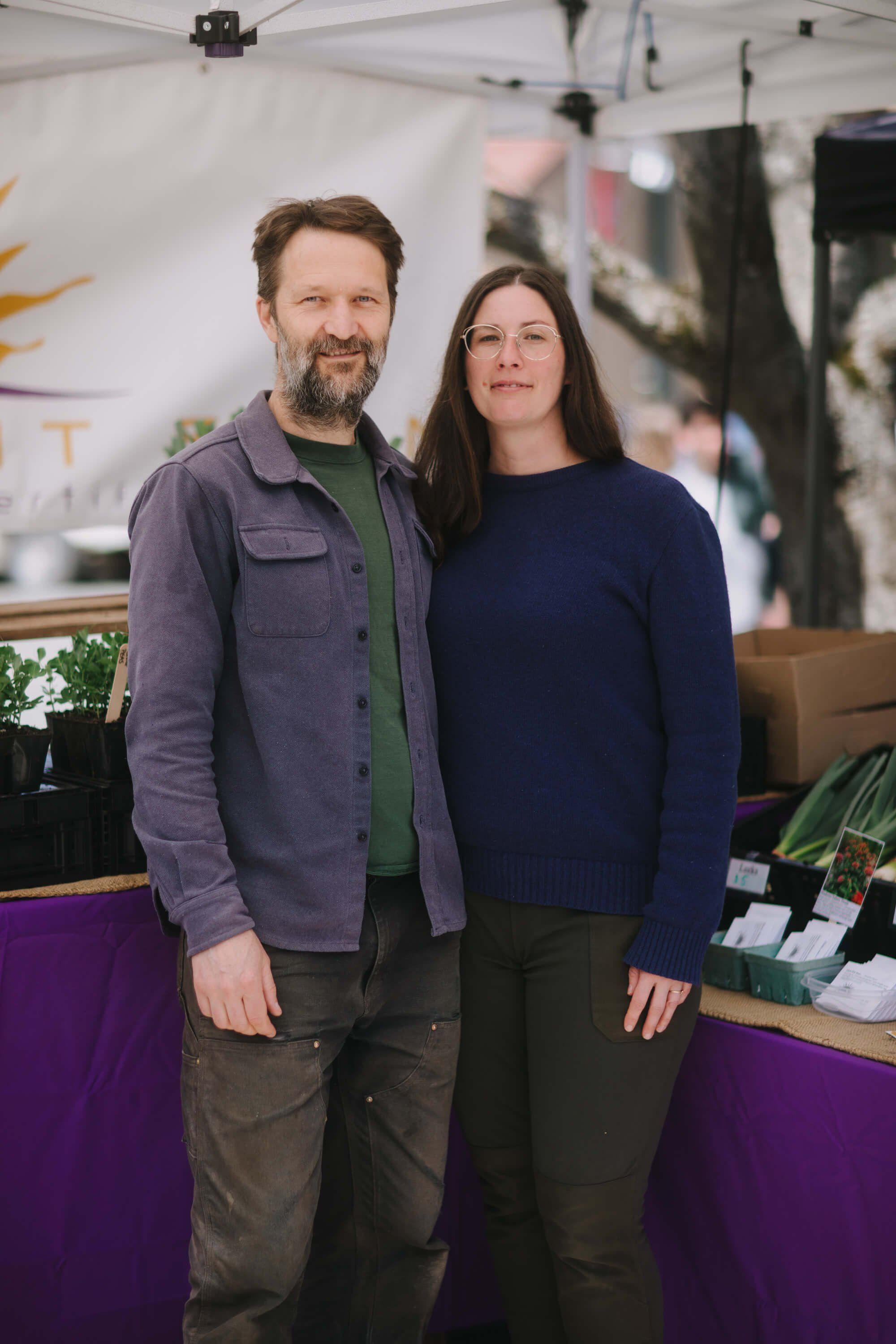 Couple standing together at Saturday Market vendor booth with purple counter, natural daylight filtering through tent canopy above