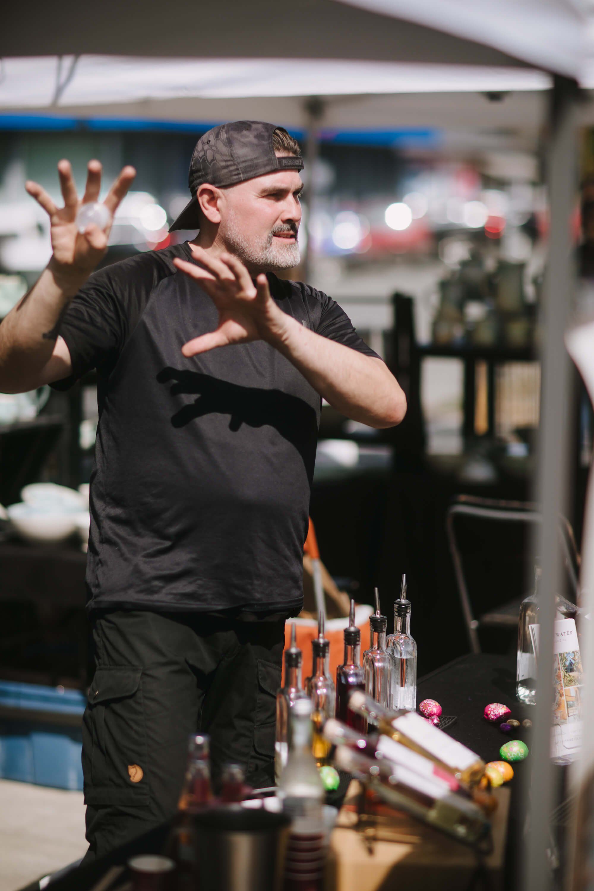Man in black shirt demonstrating at market stall with glass bottles, gesturing expressively in indoor venue