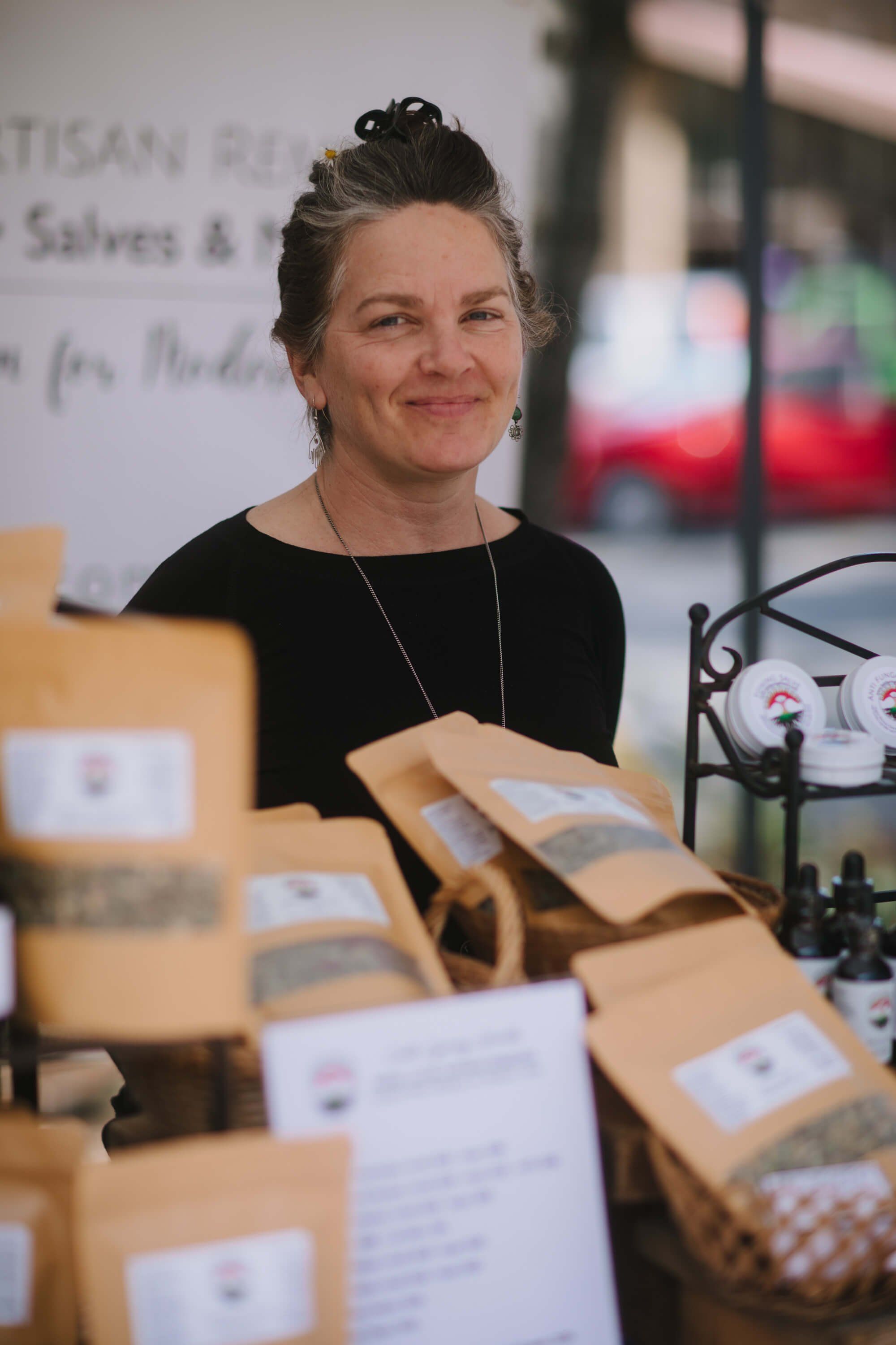 Vendor smiles beside craft packages at Saturday market stall, natural light highlighting artisan goods