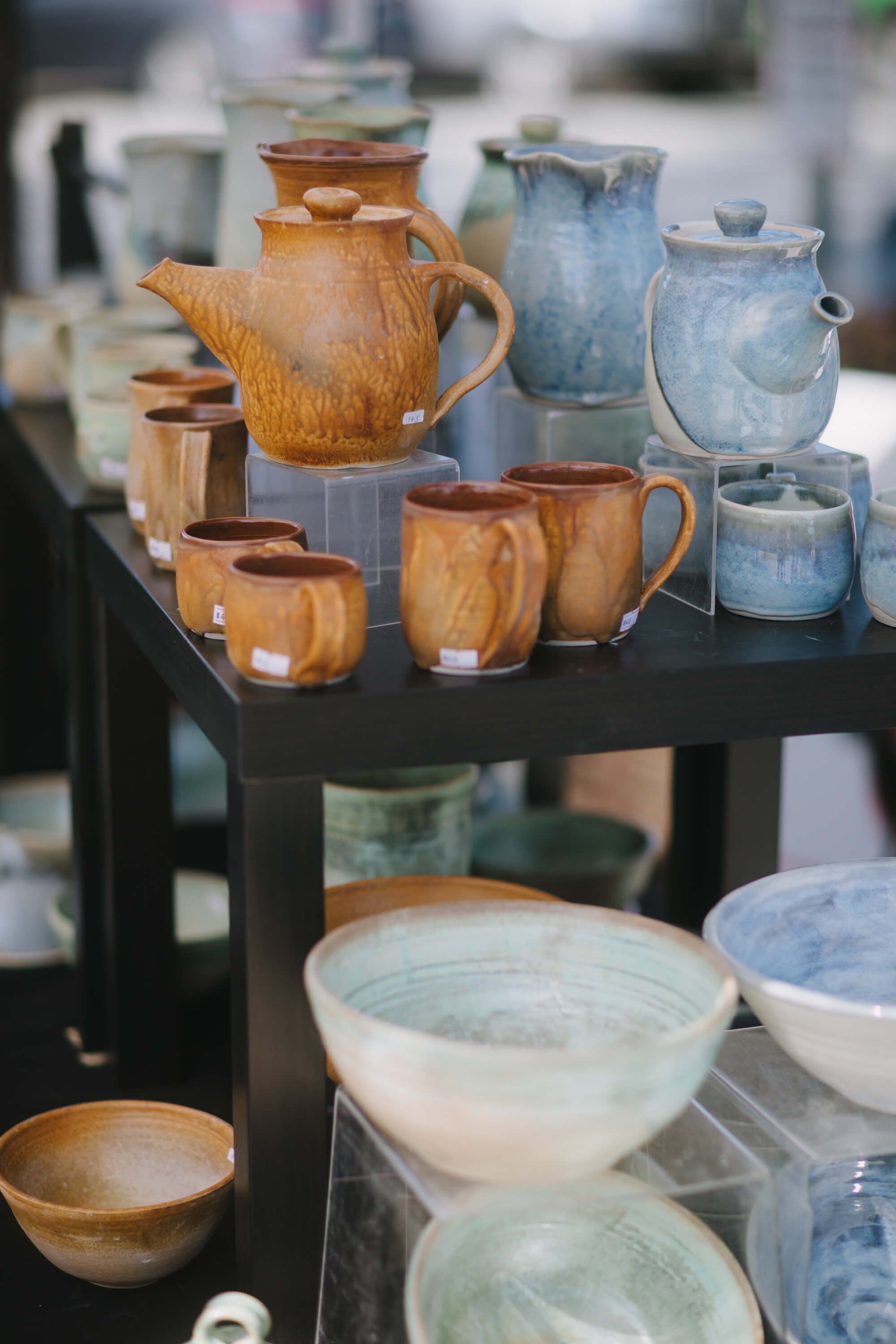 Handcrafted wooden and ceramic vessels displayed on black shelving at a market stall, featuring warm honey-toned bowls and pitchers alongside cool blue pottery