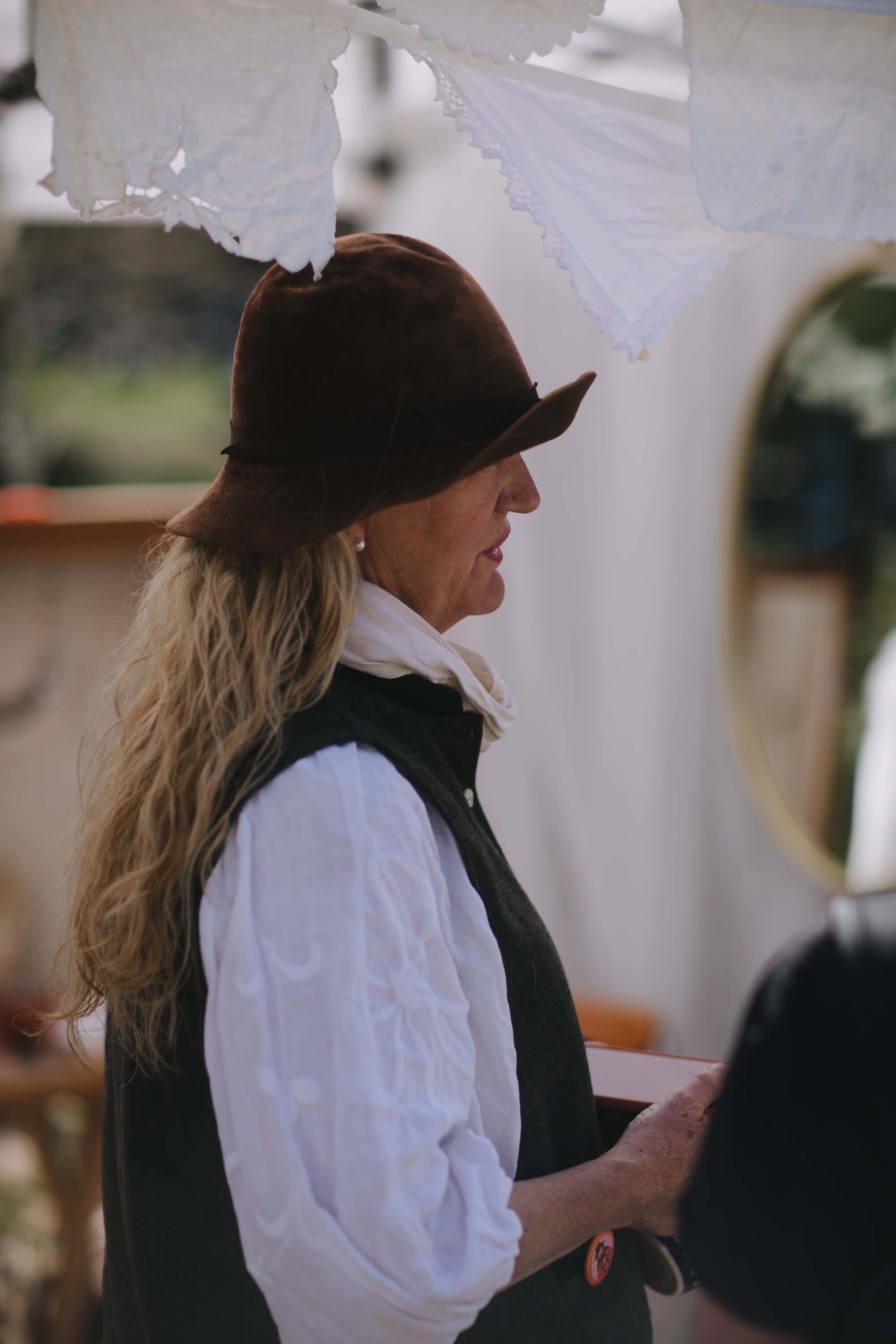 Long-haired vendor in black cowboy hat and white shirt stands at market stall beneath white fabric canopy
