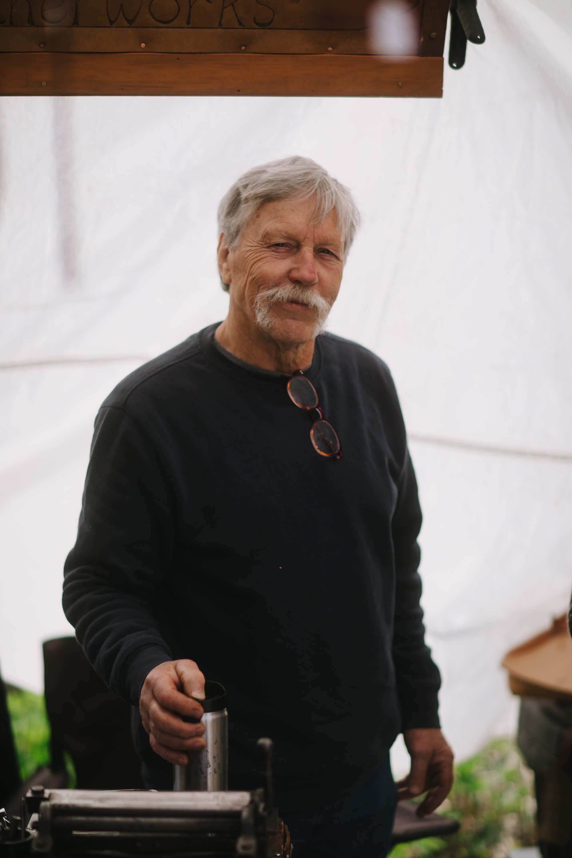 Older man with gray hair and mustache wearing black sweater and sunglasses around neck, smiling at Saturday market under white tent