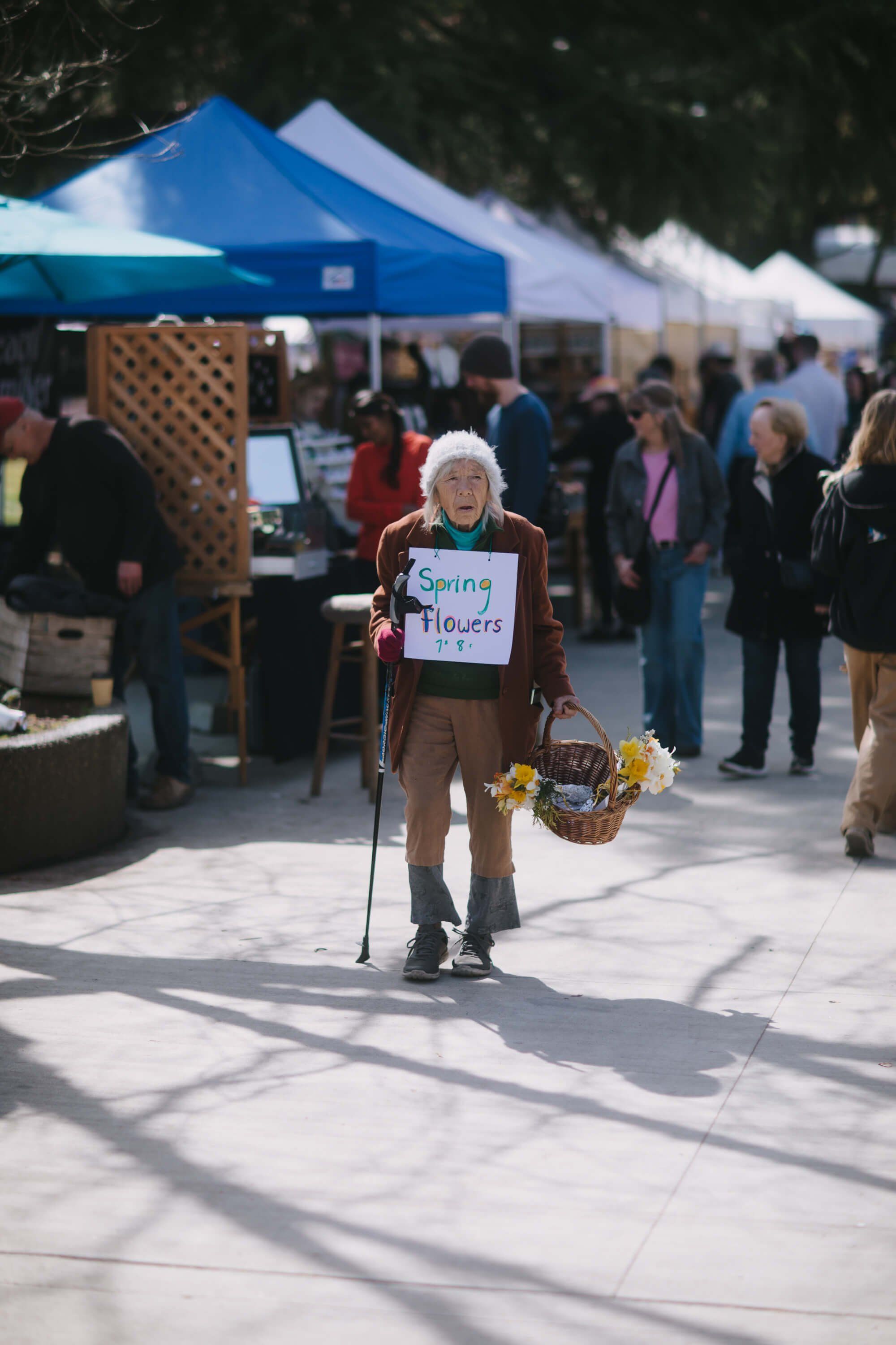 Elderly woman with white hair holding crutches and a basket of yellow flowers at a sunny outdoor market under blue and white tents