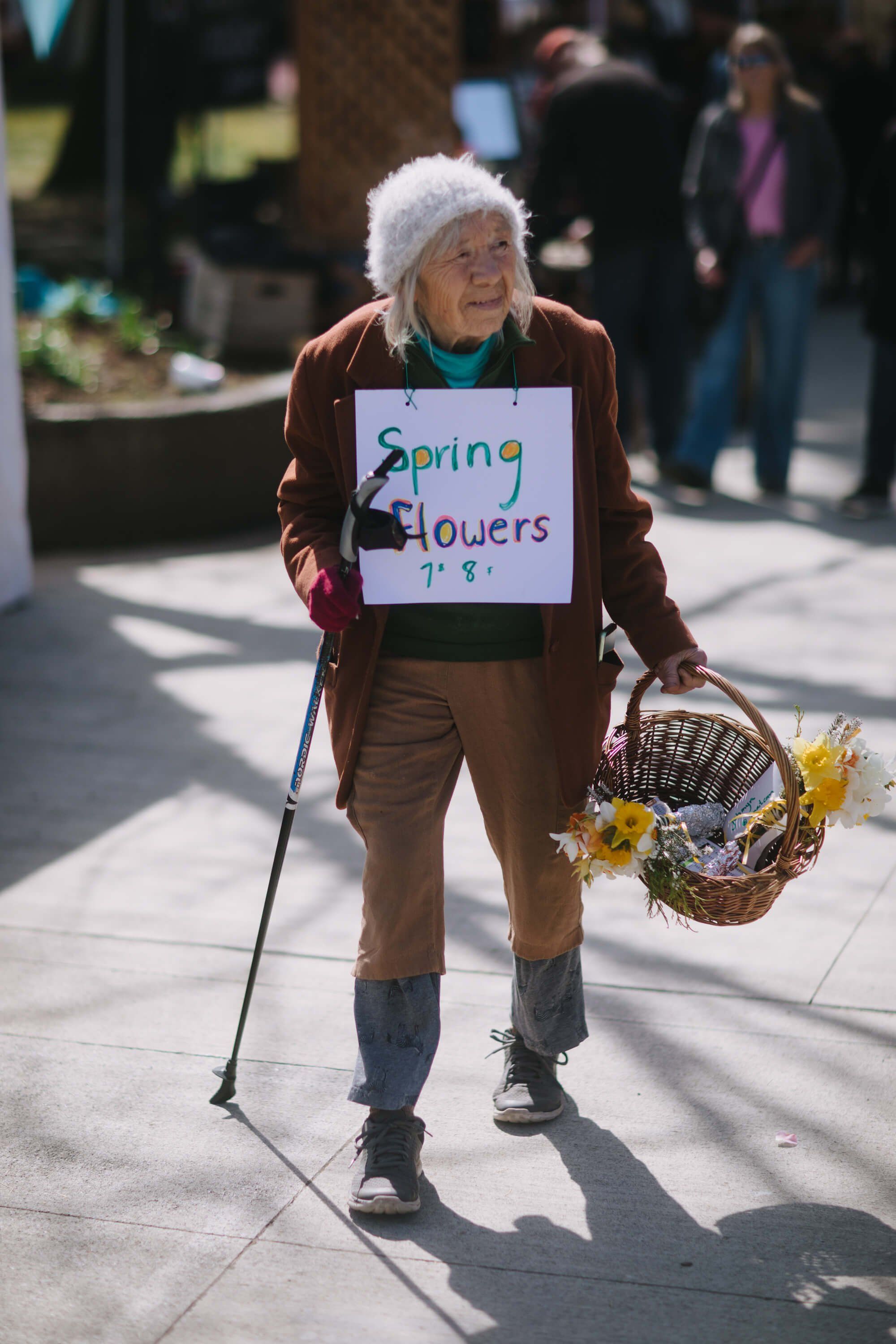 Elderly woman with white hair selling spring flowers at the Saturday Market, holding a cane and basket of yellow blooms, wearing a handmade sign