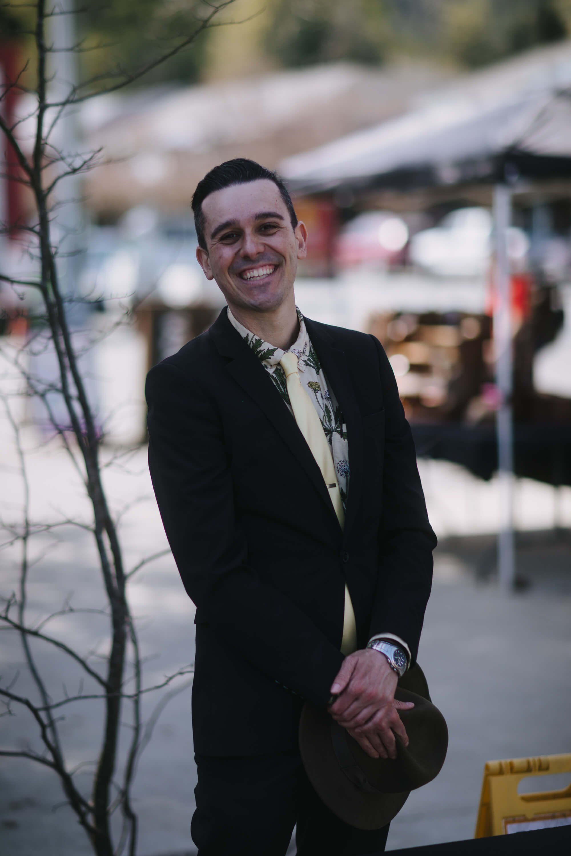 Man in black blazer smiling at Saturday Market with vendor tents and bare branches in soft focus behind him