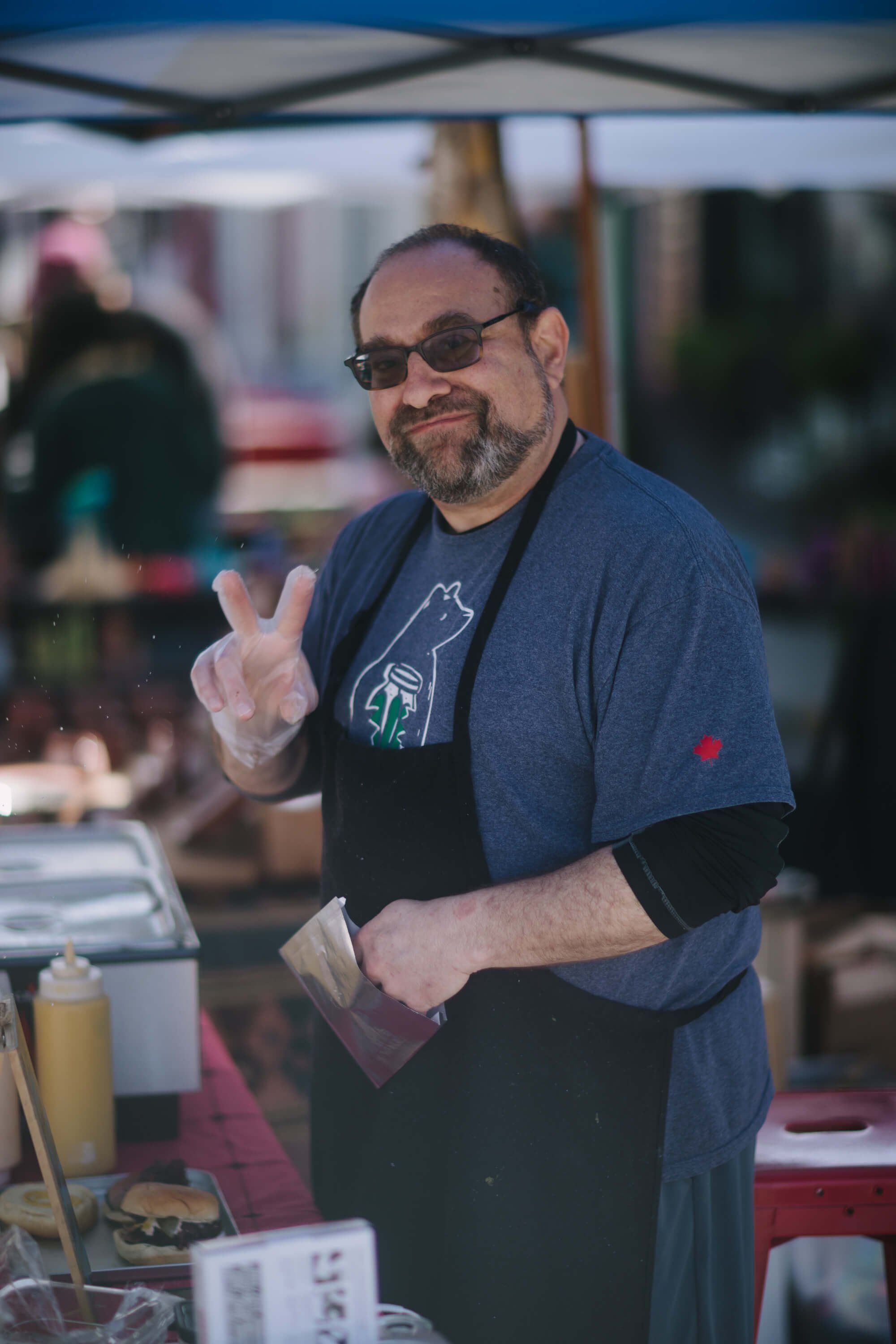 Vendor at outdoor market smiles while wearing glasses and apron, making peace sign gesture under tent canopy