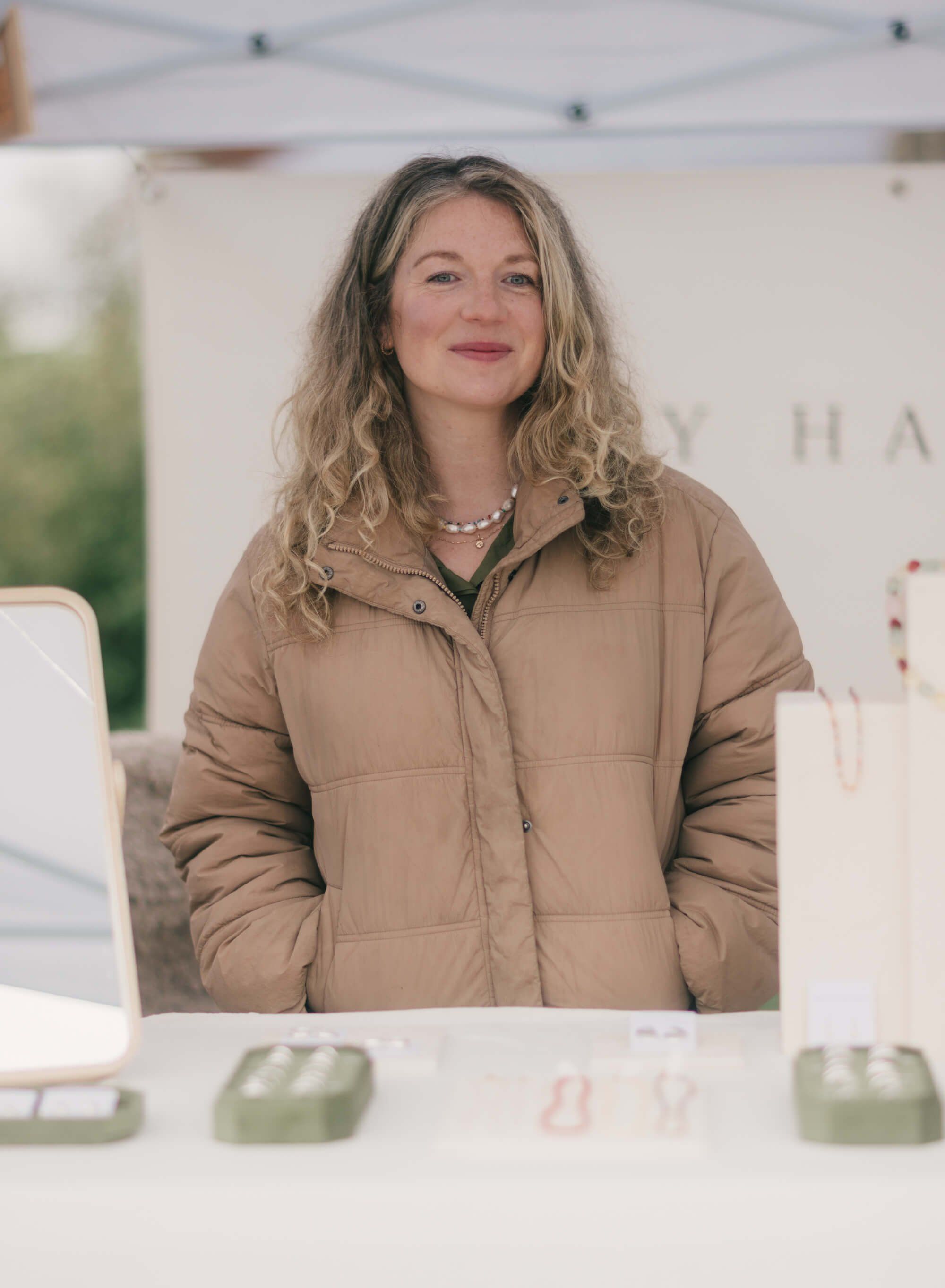 Woman in tan puffer jacket smiling at Saturday Market under white tent canopy