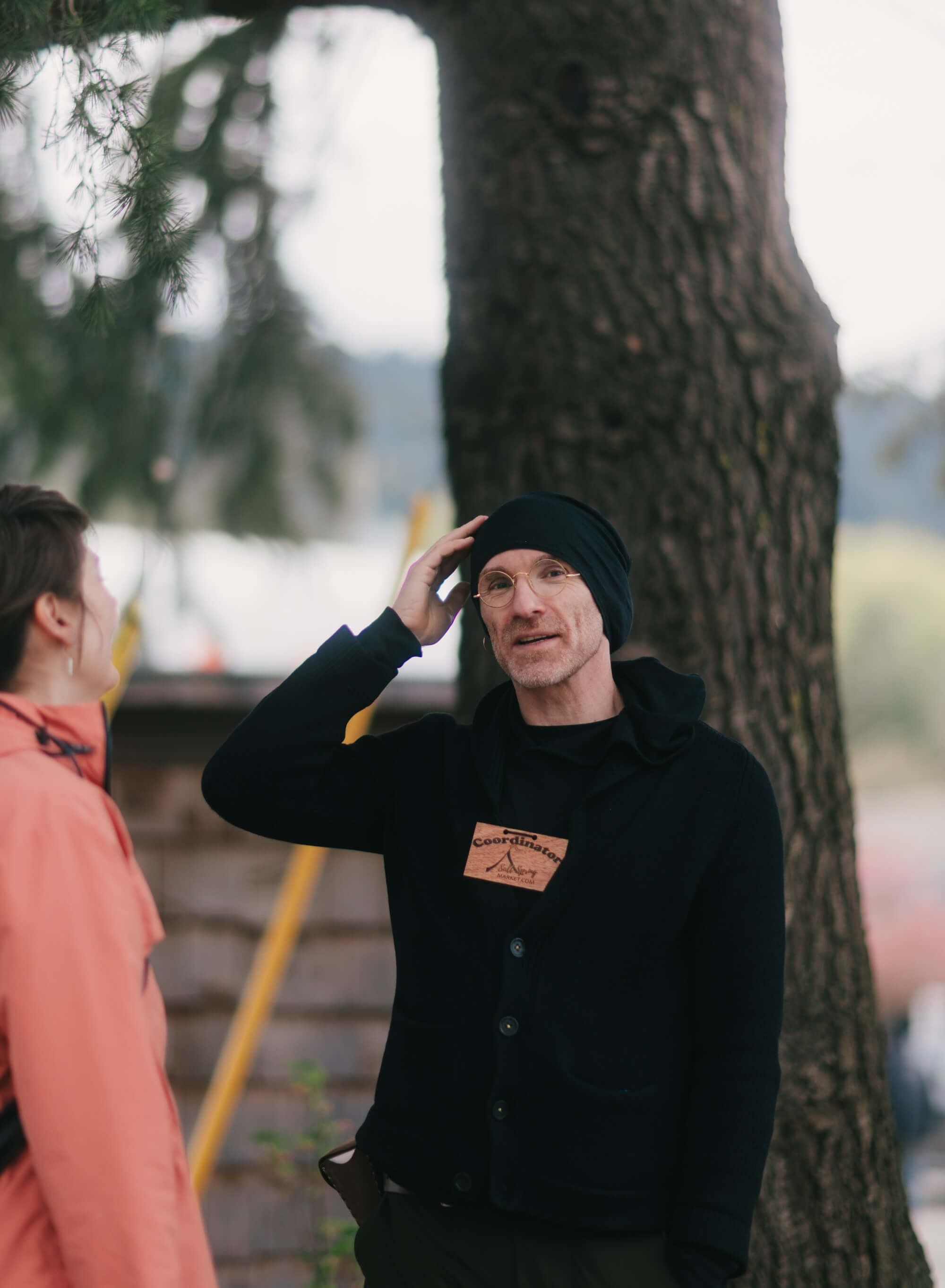 Man in black beanie and coat adjusts his hat while talking with someone in coral pink beneath a large tree at the market