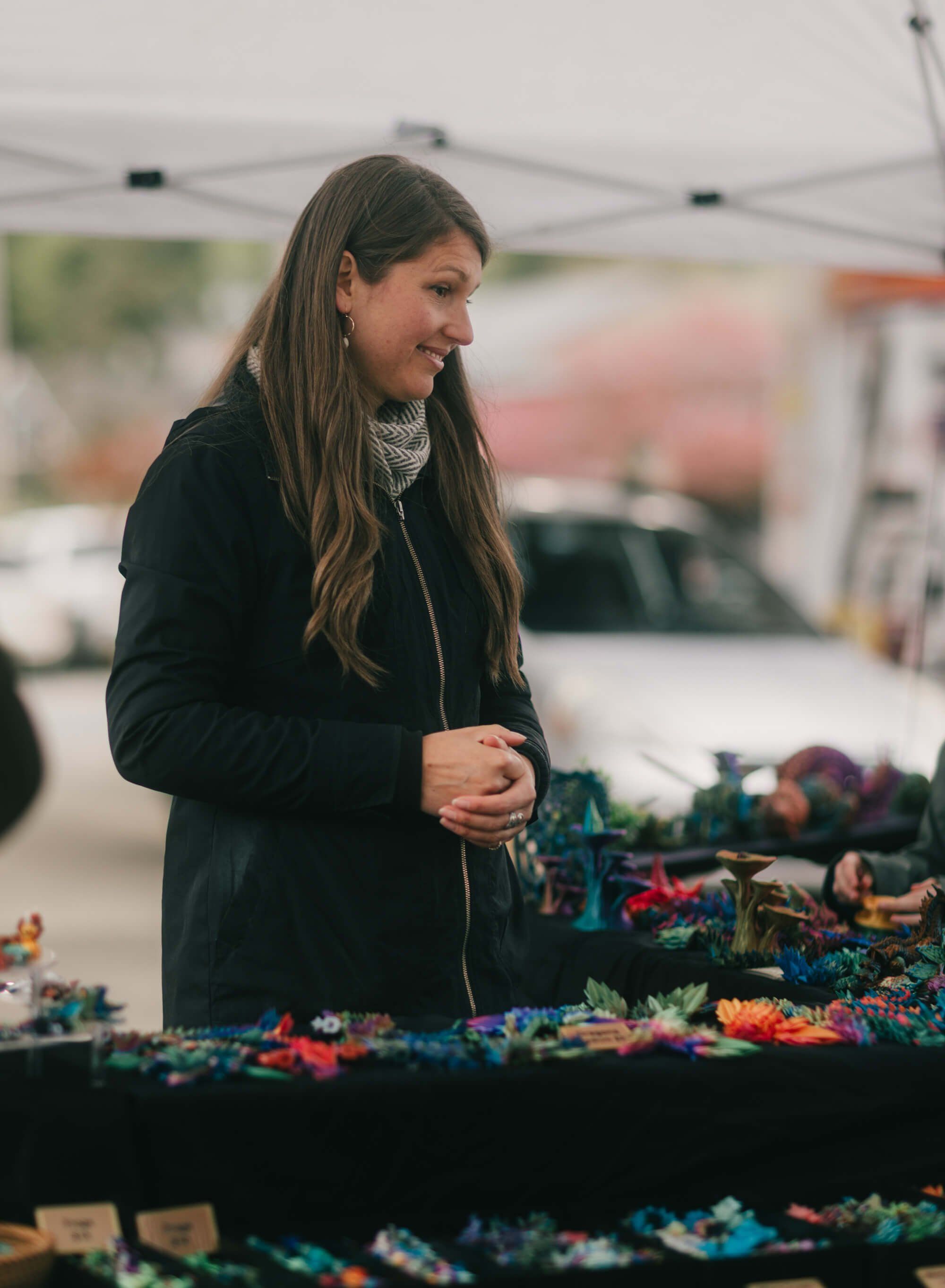 Woman in black jacket browsing colorful handmade crafts at outdoor market tent