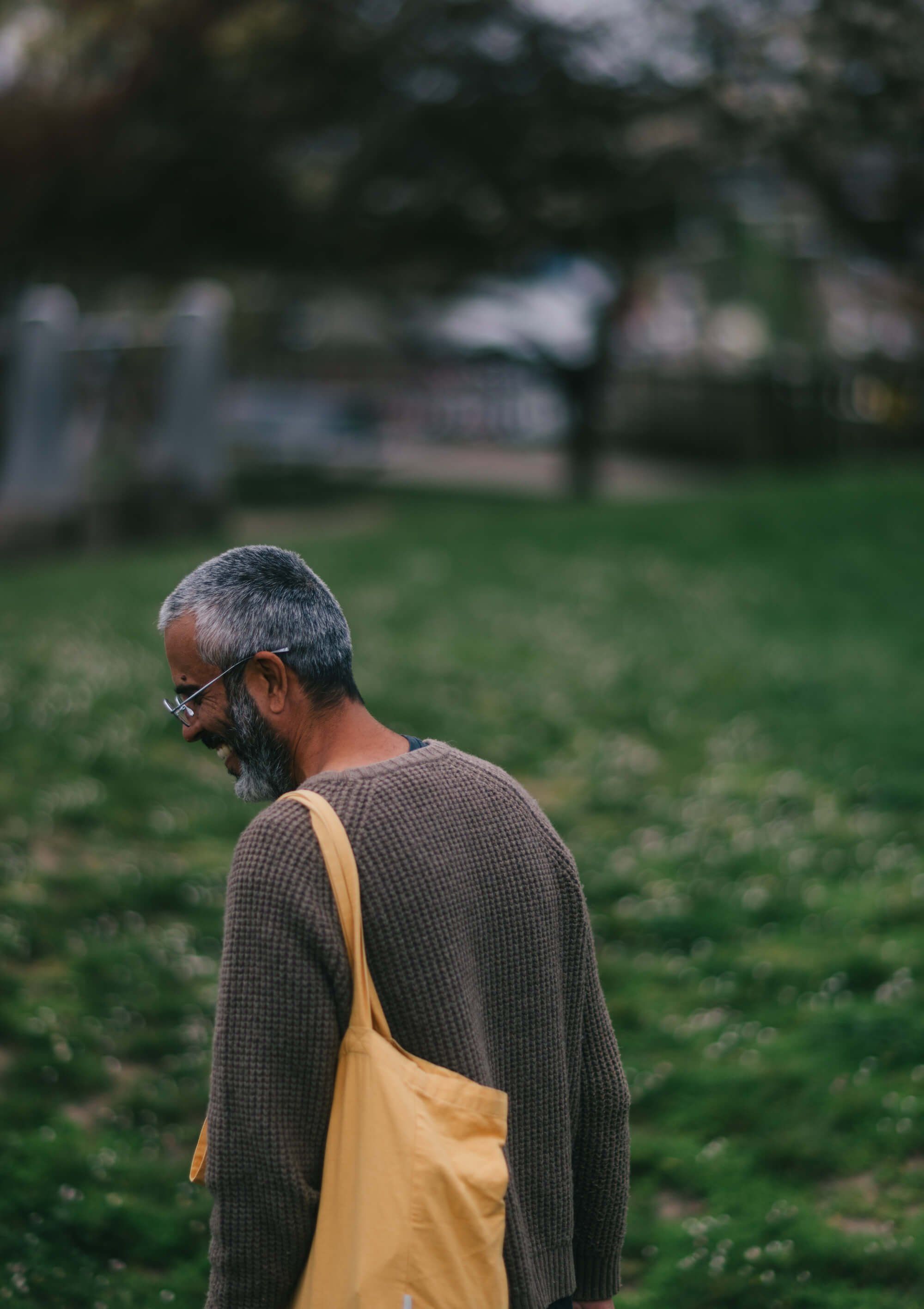 Gray-haired man in mustard tote bag and charcoal cardigan stands in sunlit park, gazing toward blurred market stalls