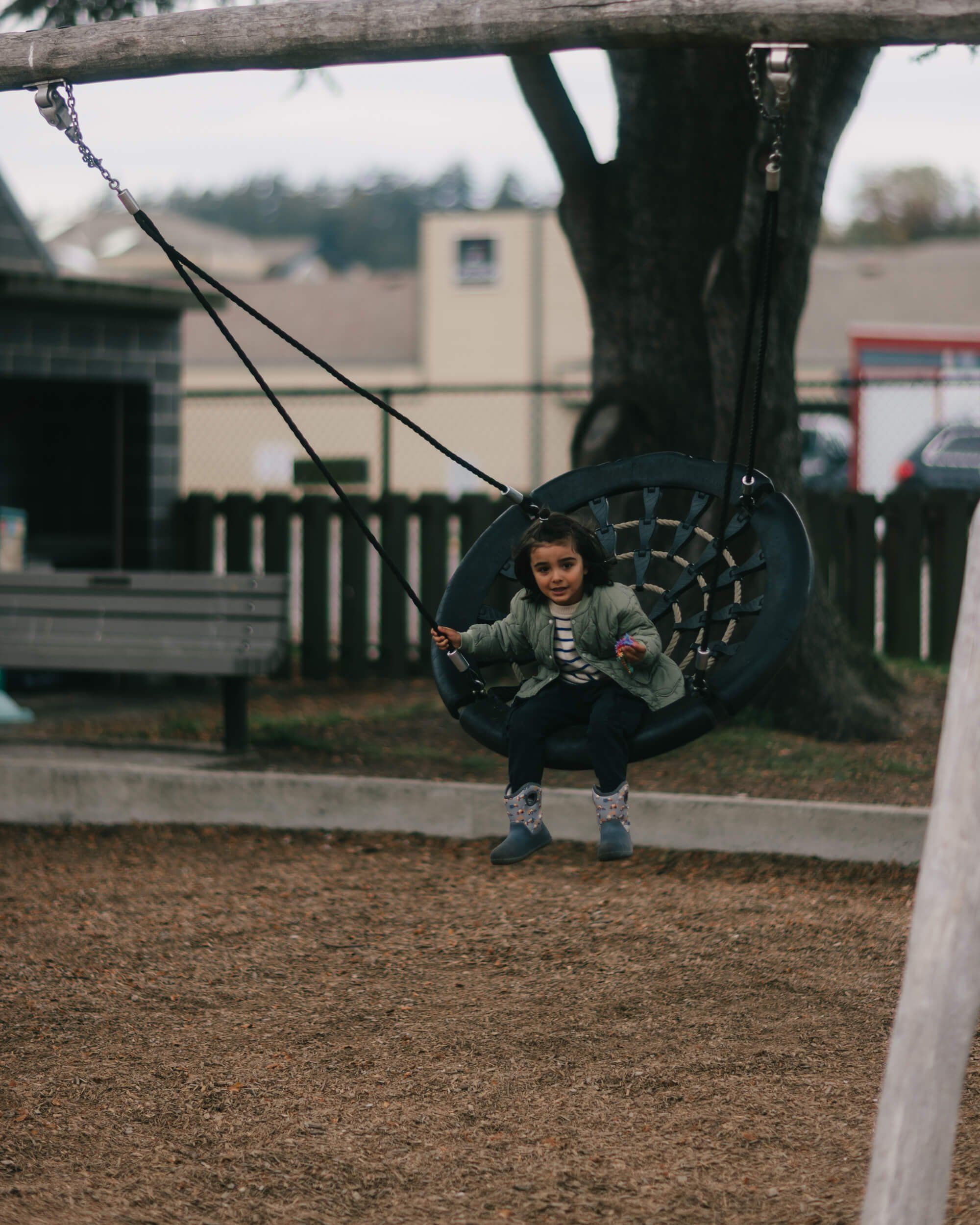 Child on tire swing beneath gnarled tree, overcast rural setting with farm buildings beyond